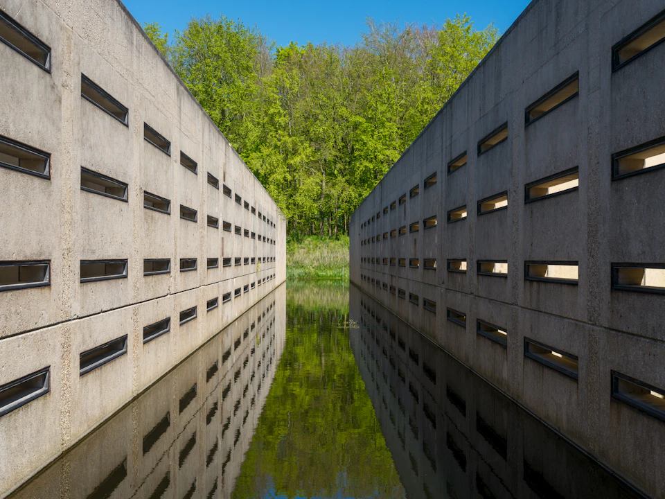 The image depicts a concrete wall with numerous rectangular windows, creating an elongated tunnel-like structure that extends into the distance. The walls are adorned with black grates on top and appear to be made from concrete or stone material. A small body of water is located at the bottom right corner of the wall, reflecting the surrounding environment. In the background, there are trees visible, adding a touch of nature to the urban setting.