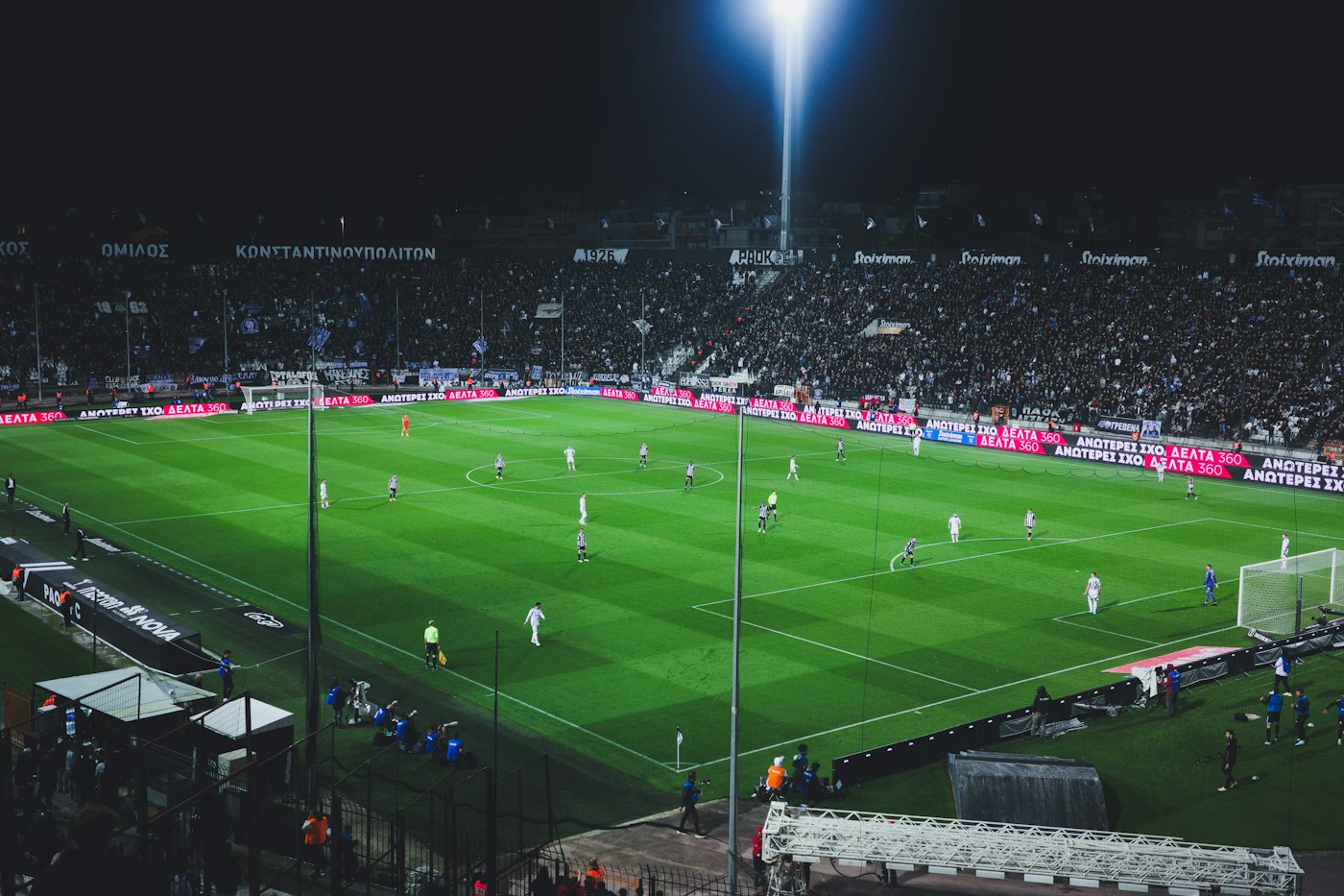Wide view of a night soccer match in a floodlit stadium with a packed stand and players spread across a green pitch