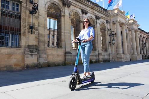 Woman rides an electric scooter near a stone building.