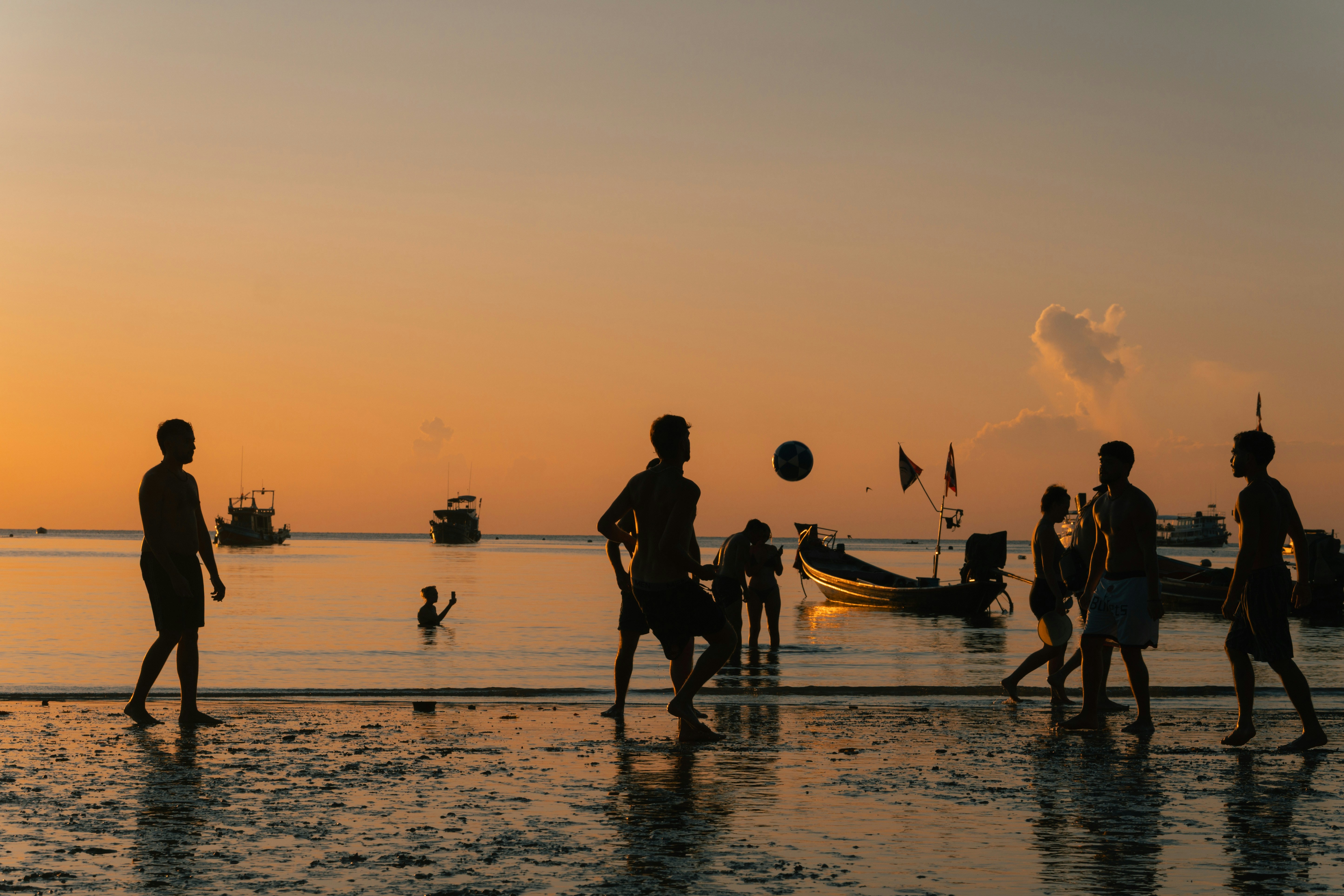 Orang-orang bermain sepak bola di pantai saat matahari terbenam.
