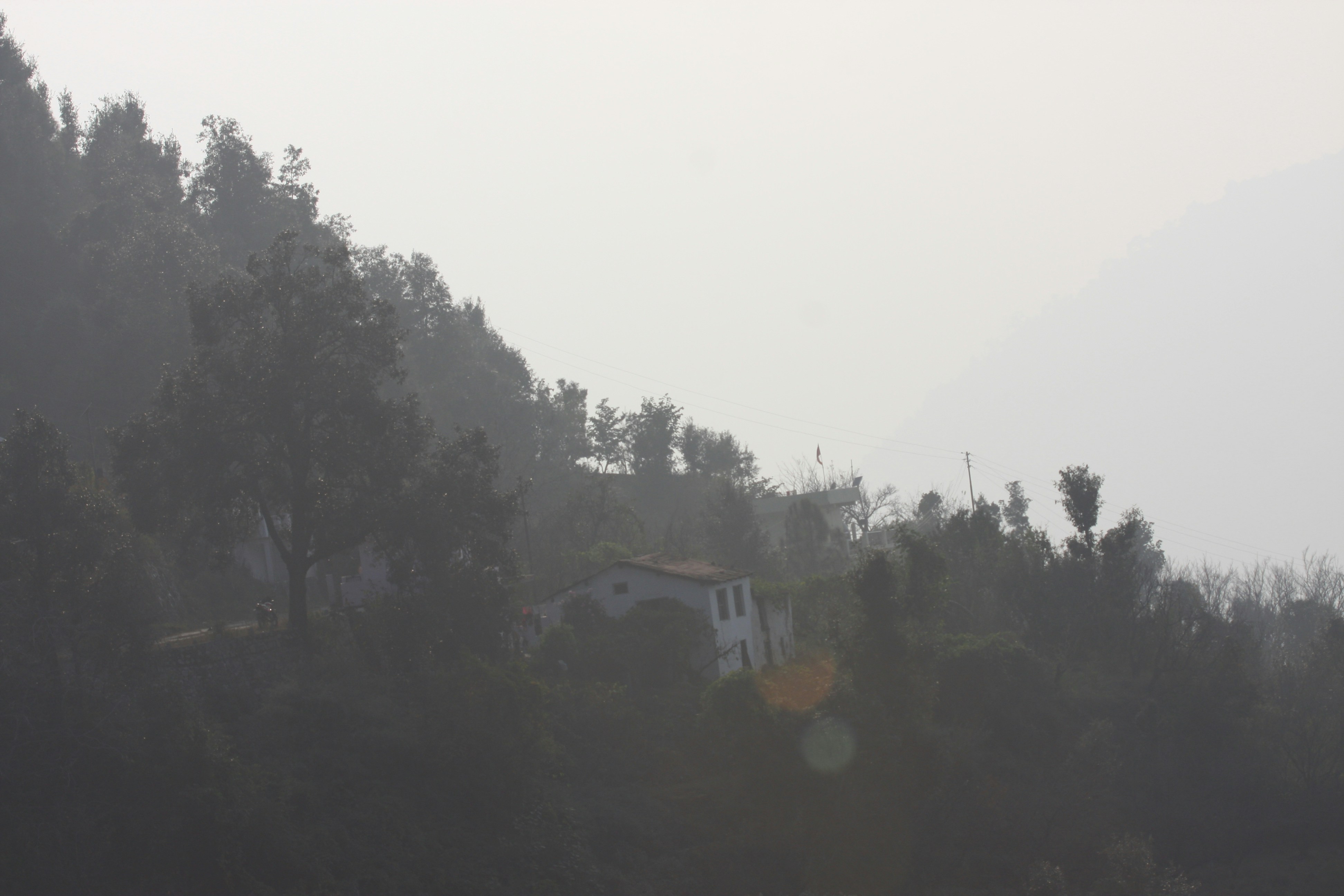 एक फैक्ट्री की चिमनी से घना धुआँ निकल रहा है, जिसके सामने एक हरियाली वाला गाँव और लोग दिख रहे हैं, जो प्रदूषण से प्रभावित लग रहे हैं।