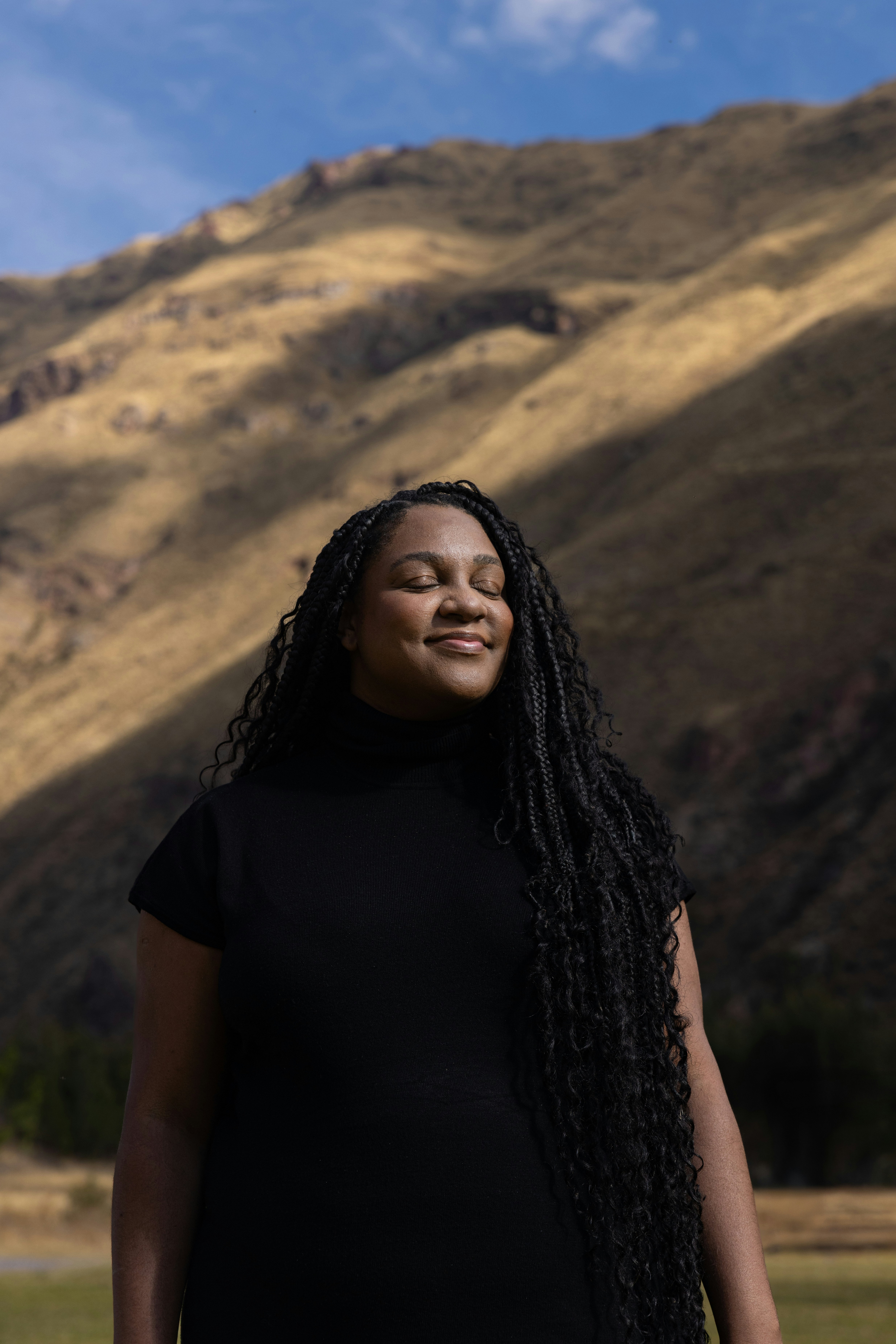 Woman smiles with eyes closed near a mountain.