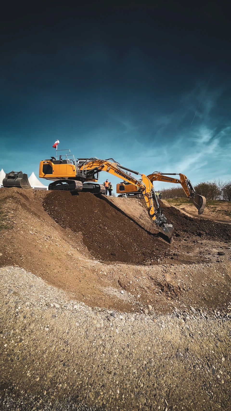 Two yellow excavators, one digging, the other stationary, at a construction site with multiple piles