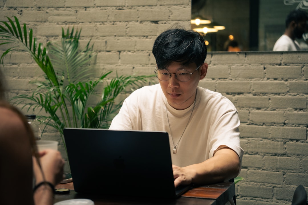 Man works on a laptop at a cafe., An individual, who appears to be an Asian man wearing glasses, is working on a laptop at a cafe. The setting suggests a relaxed indoor environment.