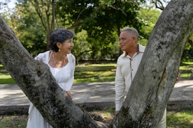 Couple smiles at each other behind two trees.