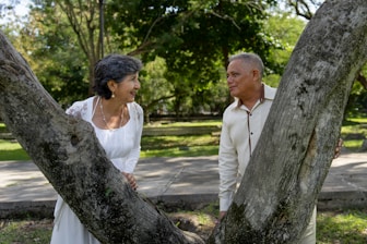 Couple smiles at each other behind two trees.