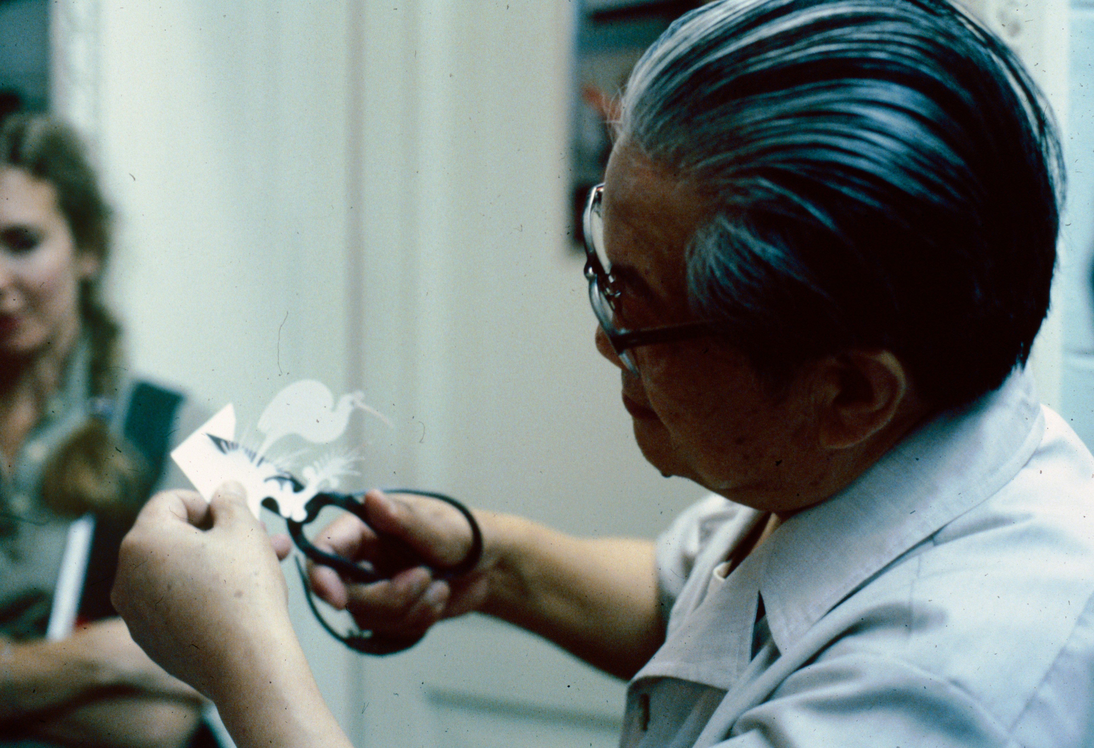 Man carefully cutting paper, watched by a woman.