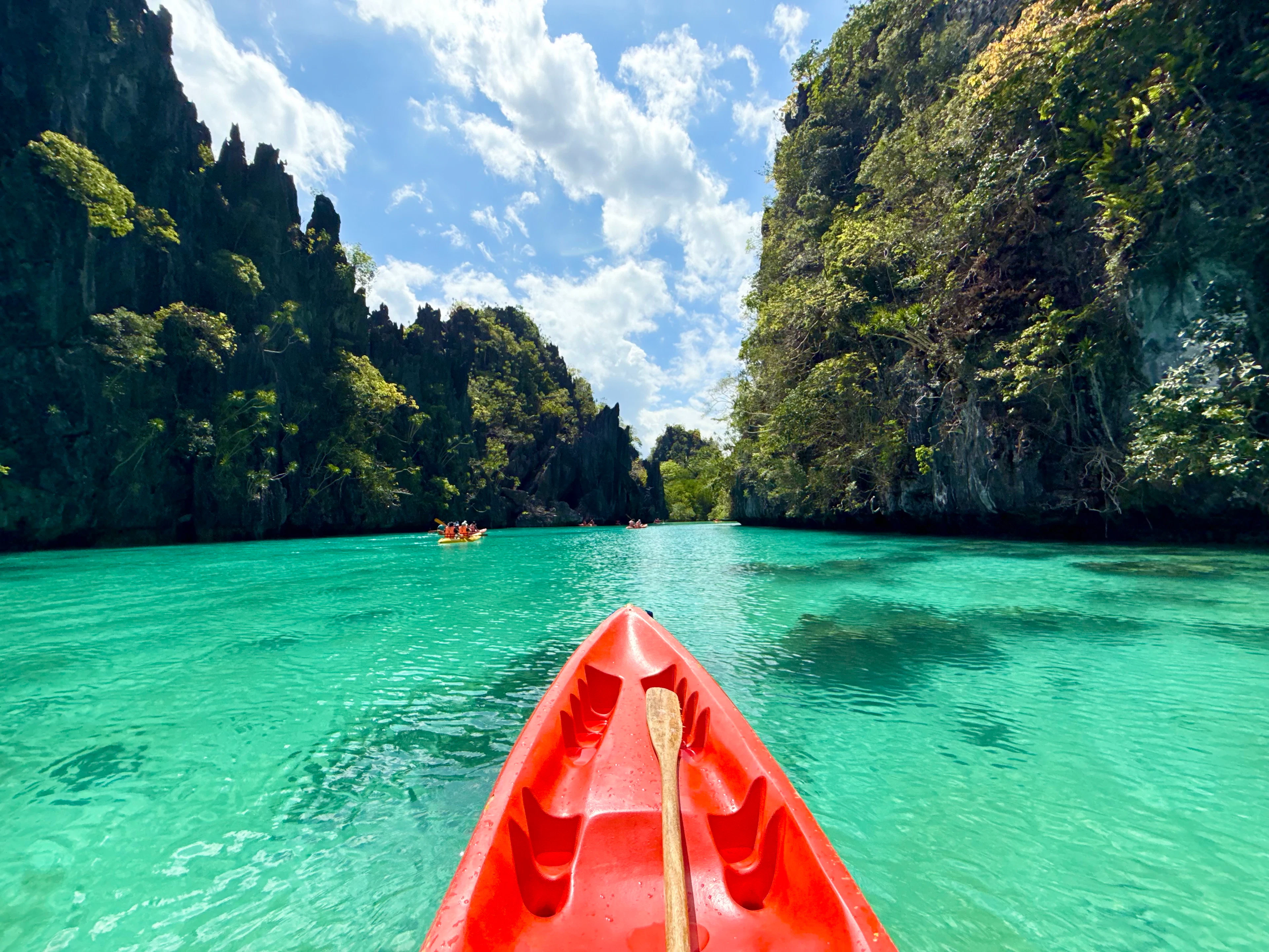 The bow of a red kayak on turquoise water between the cliffs of El Nido