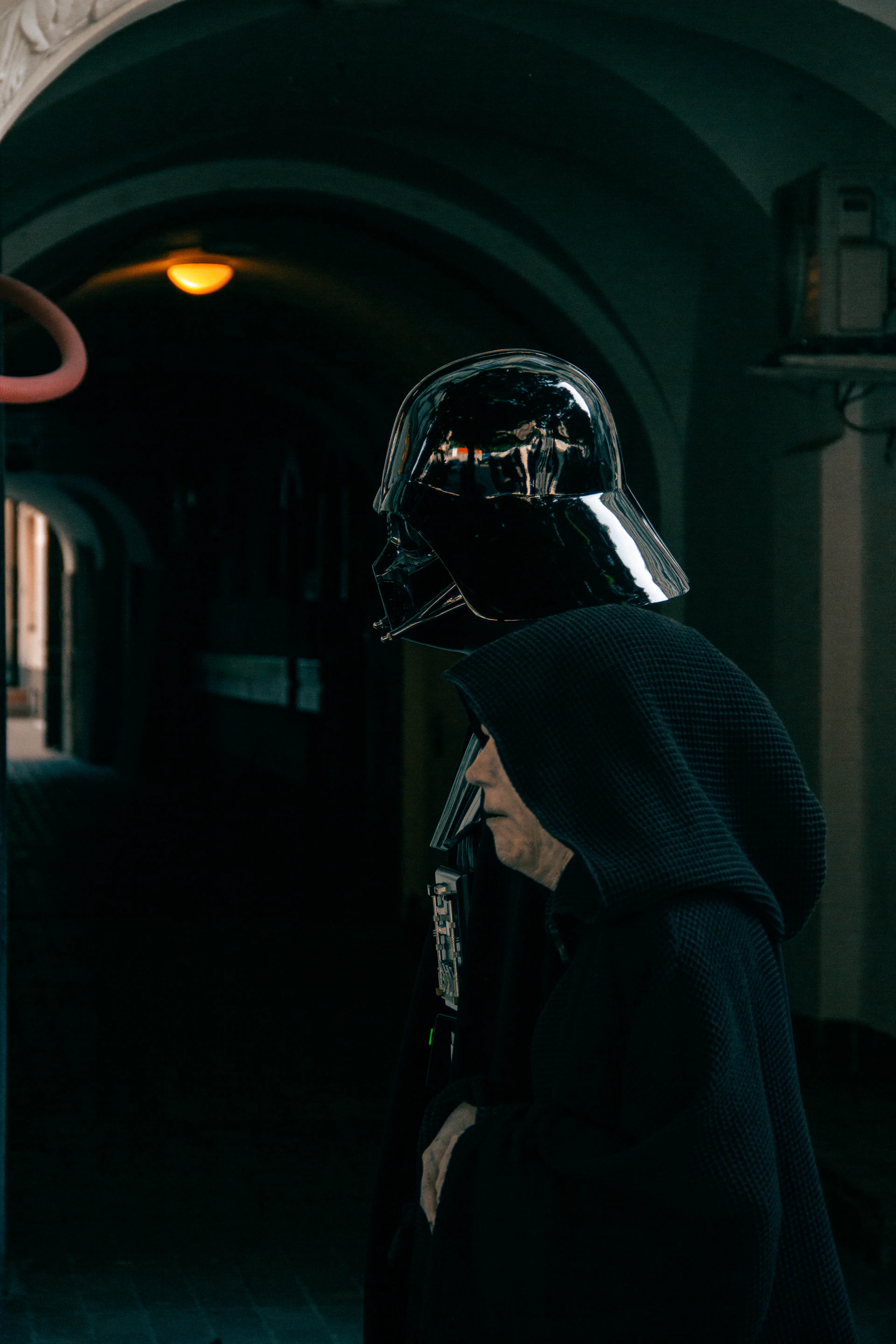 Darth vader walks through a dark, arched passage.