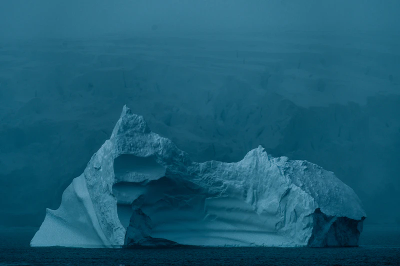 A blue and white iceberg floating in deep dark Antarctic waters under a soft sky