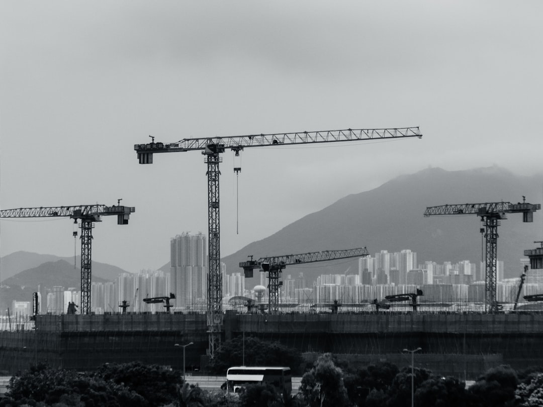 Construction cranes populate the cityscape., The skyline is punctuated by construction cranes at a major hospital site in Kai Tak, Hong Kong. In the distance, clusters of high-rise buildings across Victoria Harbour on Hong Kong Island East are partially veiled in mist. In the foreground, a double-decker bus glides along the highway, capturing the dynamic essence of urban life.