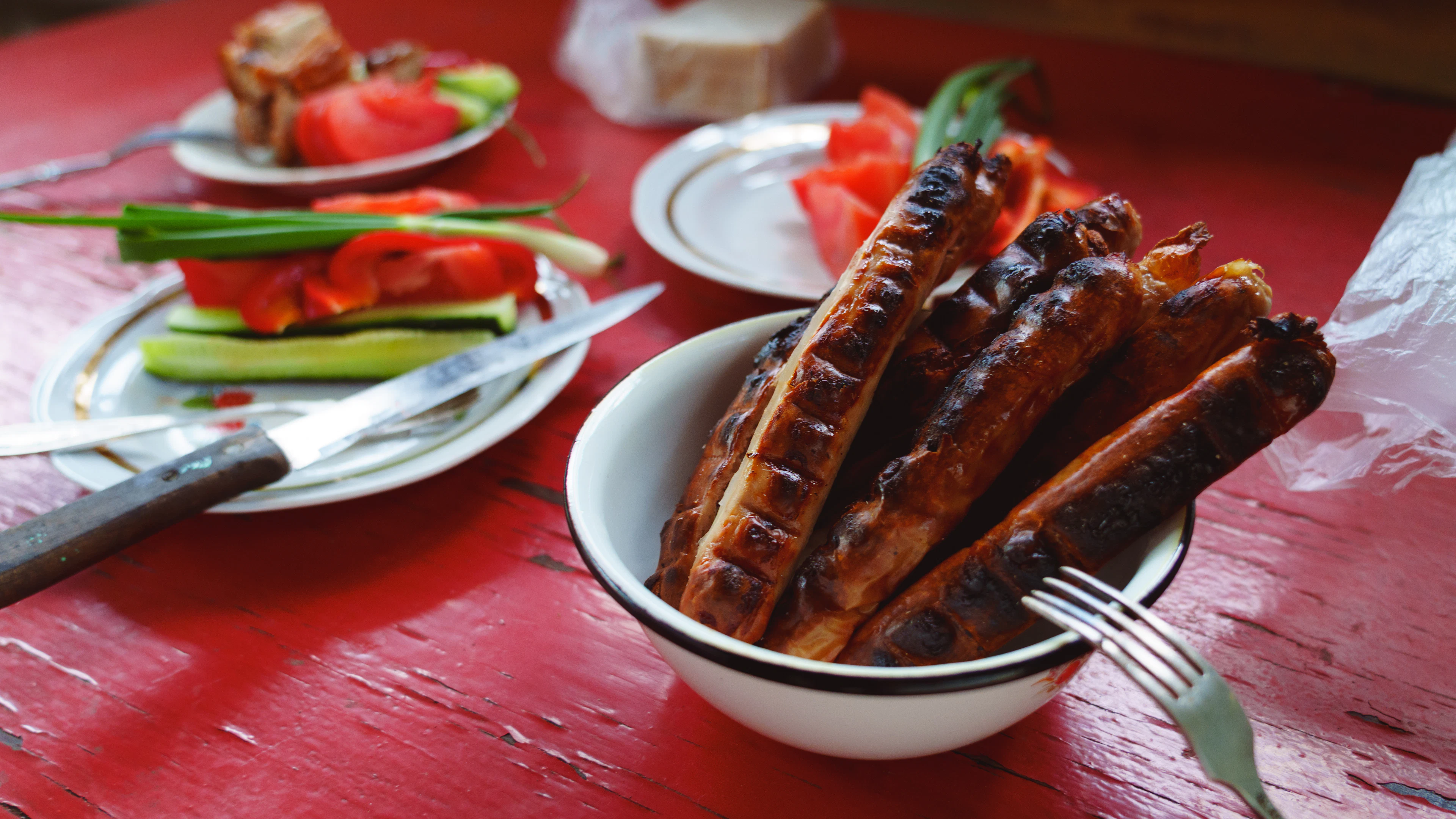 Charred grilled meats piled in a bowl on a red table, salad and roasted peppers beside them