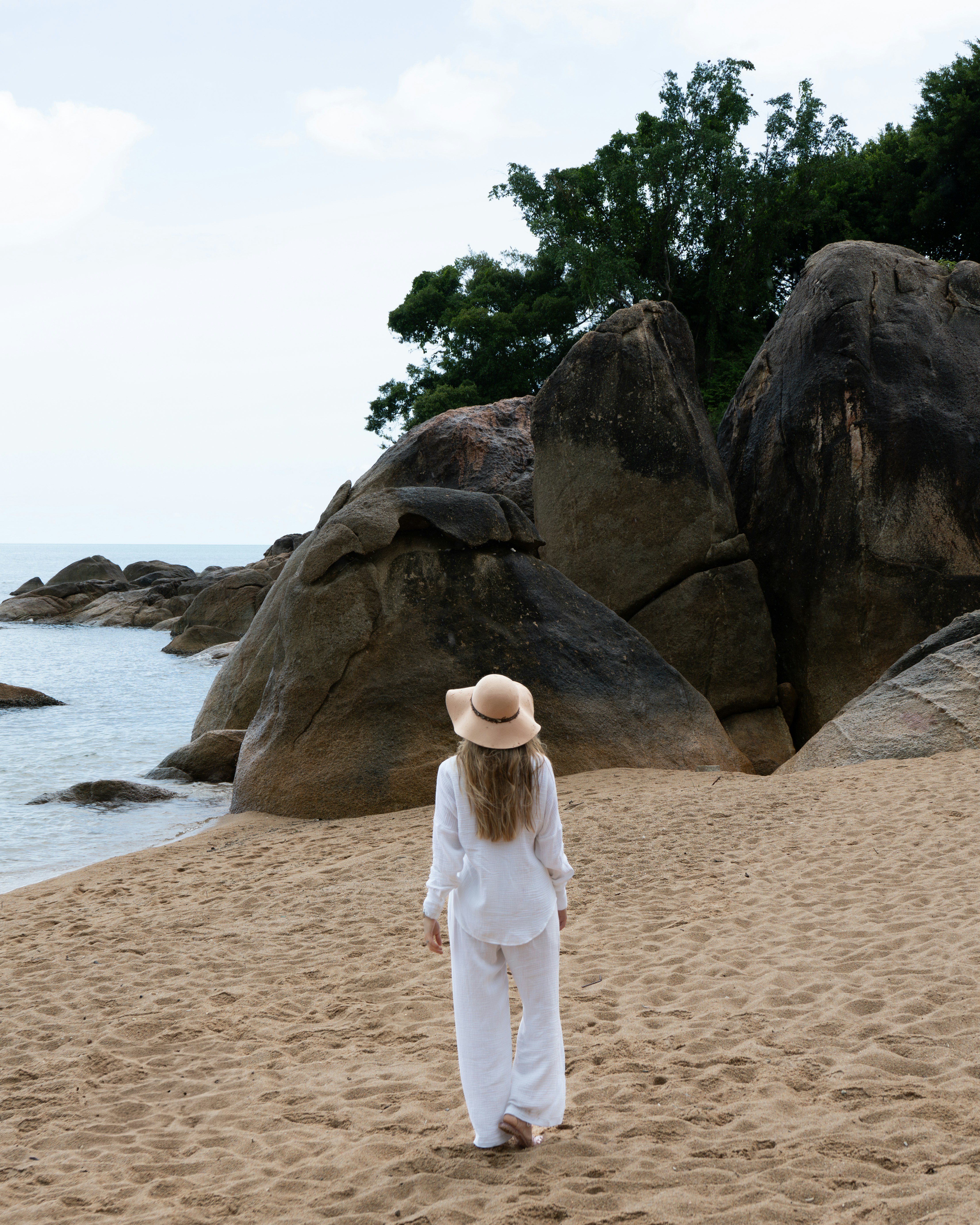 A woman walks toward rocks on a sandy beach.