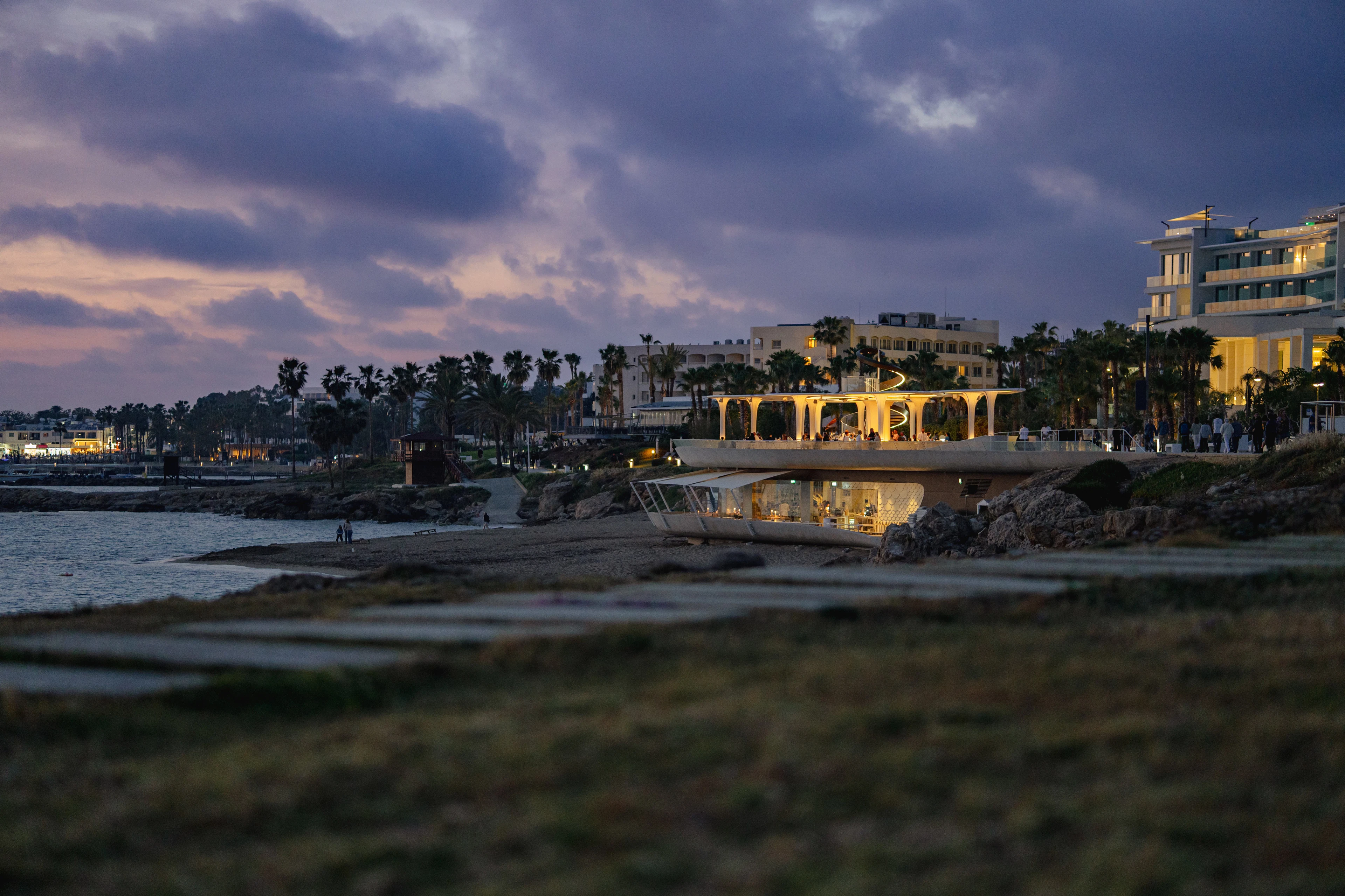 The Kato Paphos seafront at dusk under a violet sky, a lit beach pavilion glowing on the rocks and the white blocks of the seafront hotels standing among palm trees behind the shore