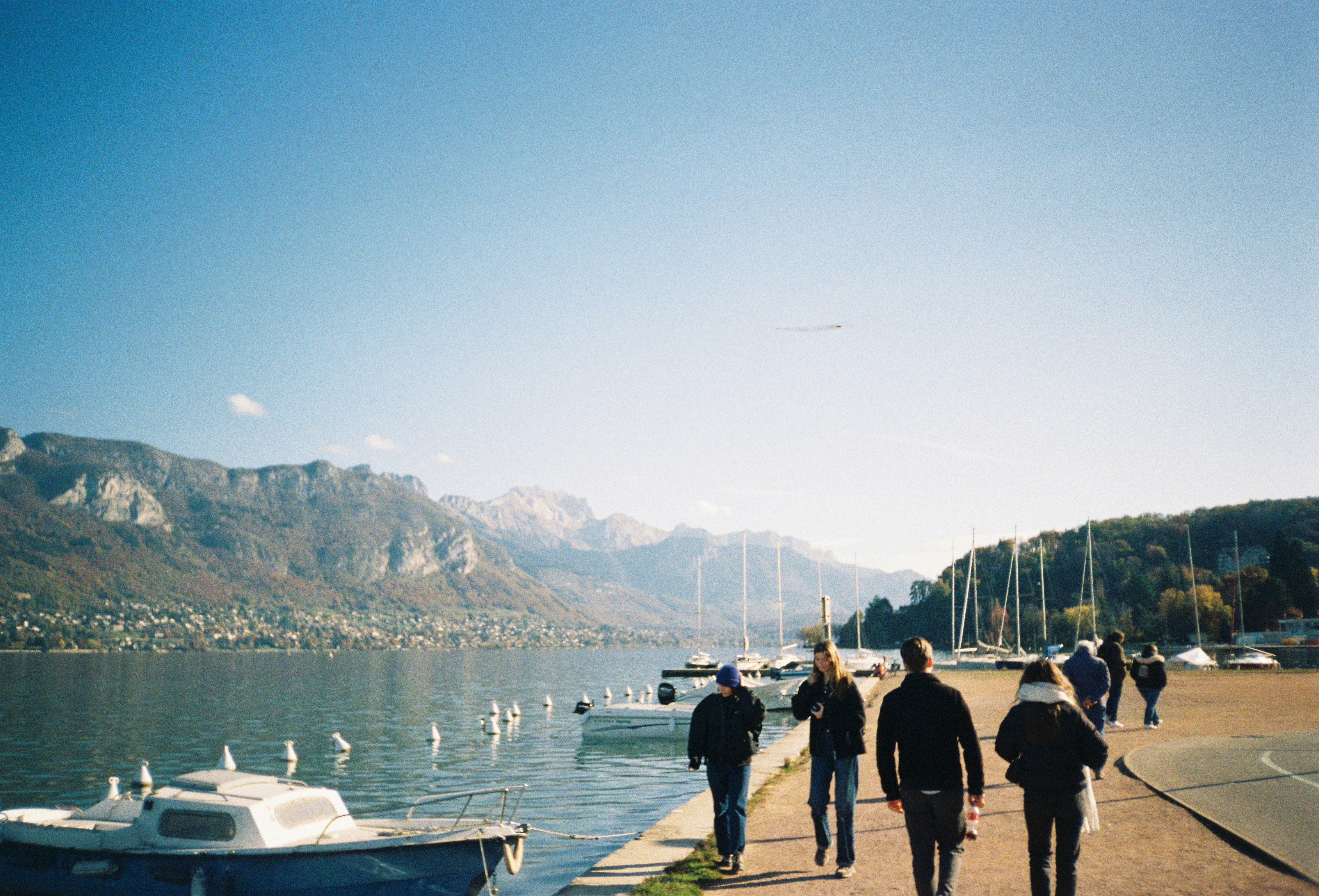 People walk along a lake with mountains in the background.