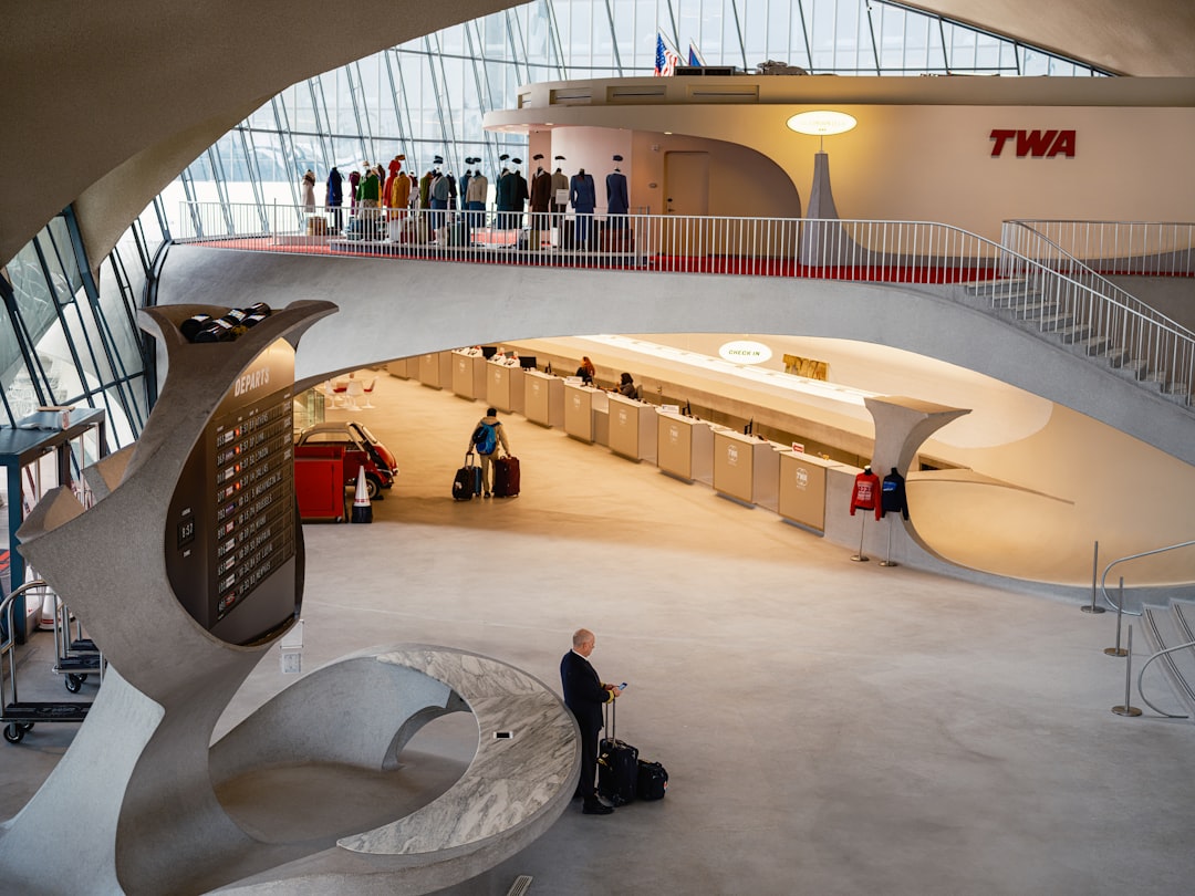 Inside a retro-styled airport terminal., Inside the TWA Hotel Flight Centre at JFK Airport in New York City. A pilot stands near the departures board as travellers walk to check in. TWA uniforms and mid-century, the scene. design enhance