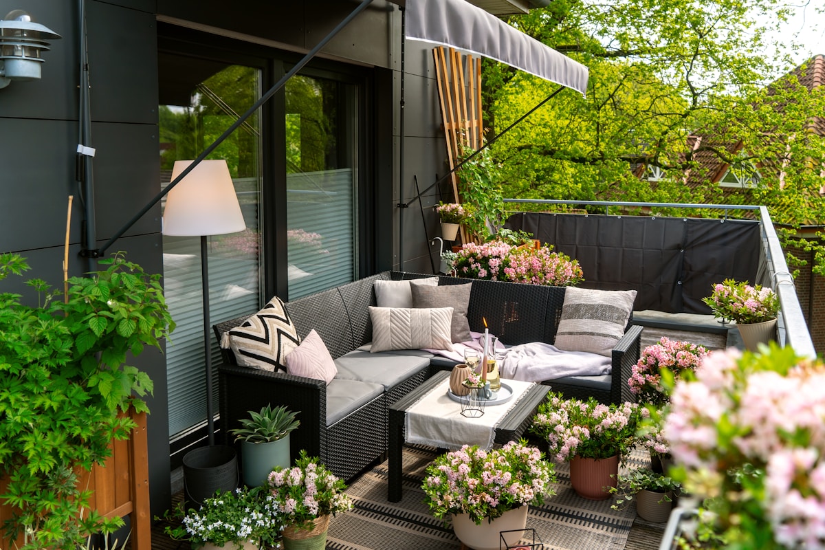 Cozy apartment balcony with rattan sectional sofa, bright pink potted flowers, bamboo screen, and a retractable grey awning