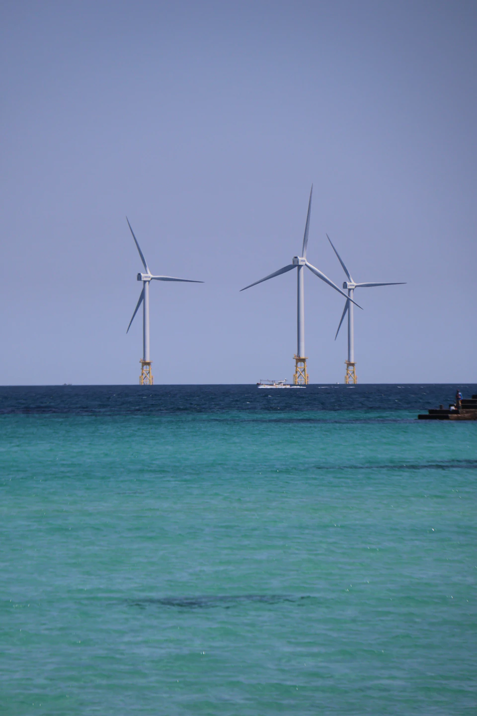 Three wind turbines float on calm ocean water with a small boat nearby