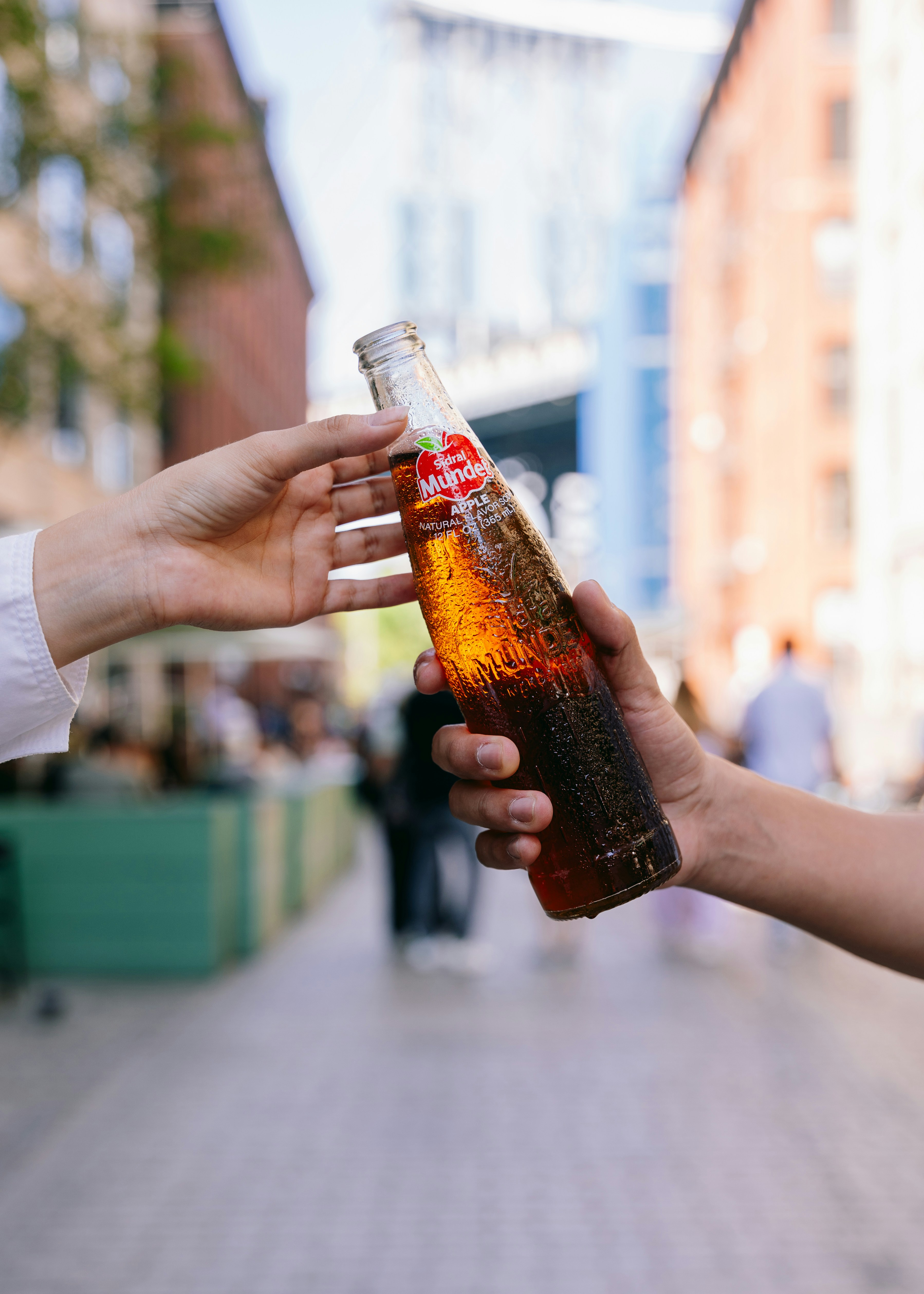 Hands passing a bottled beverage outdoors.
