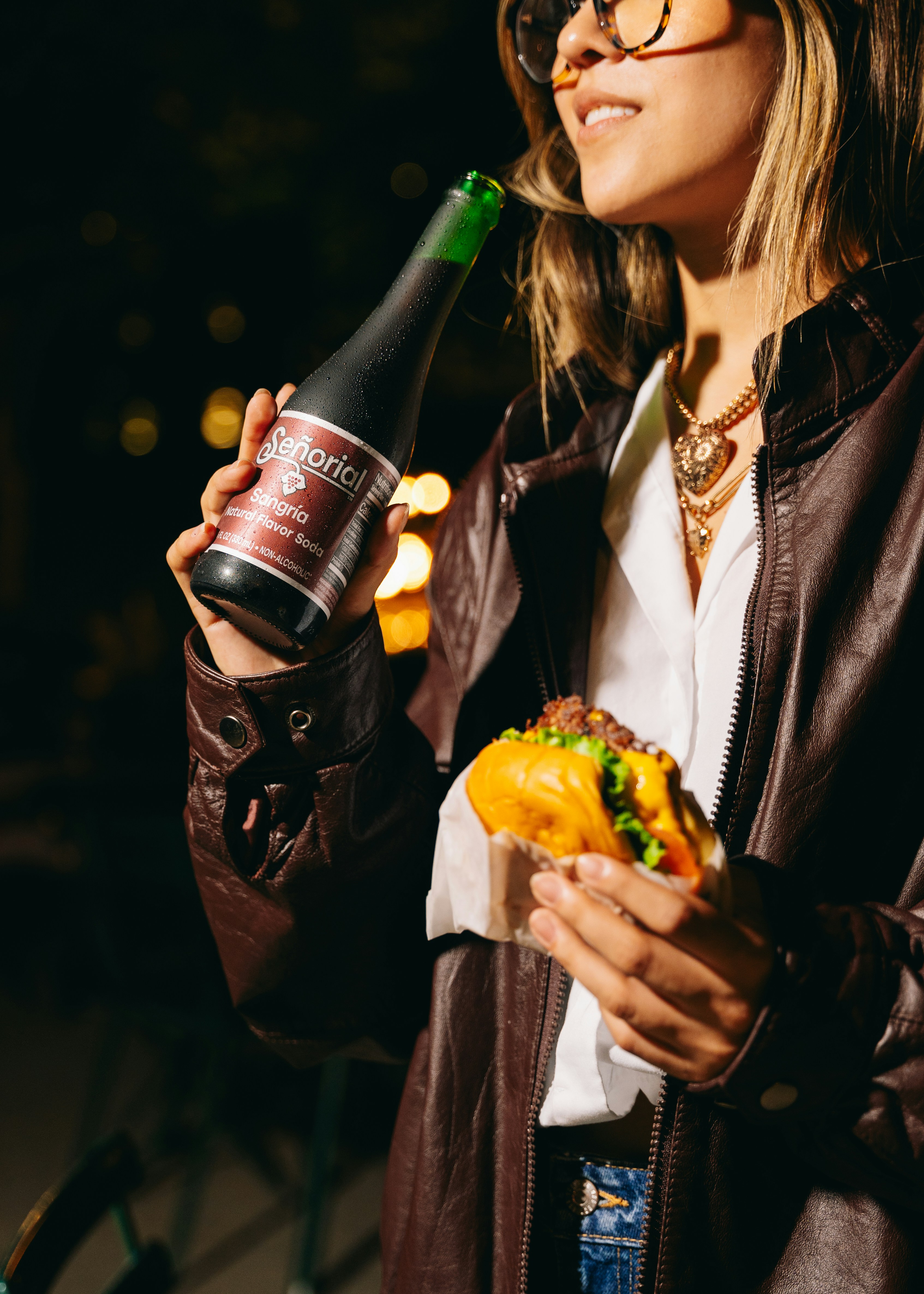 Woman enjoys a drink and taco outdoors at night.