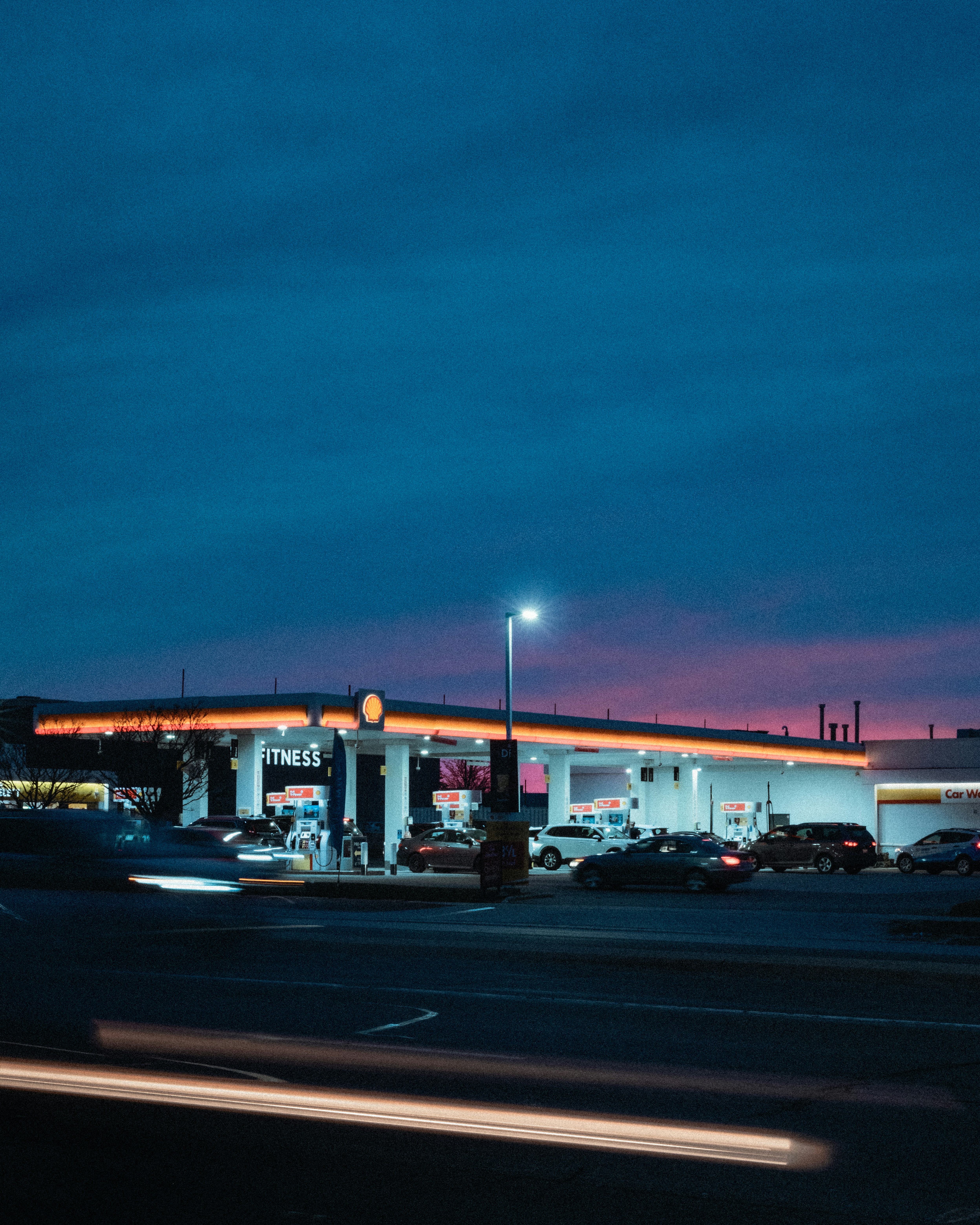 A cinematic view of urban life in Oakville as dusk settles over a glowing Shell gas station. Neon lights pop against the fading sky, and car headlights streak past, capturing a fleeting everyday scene.