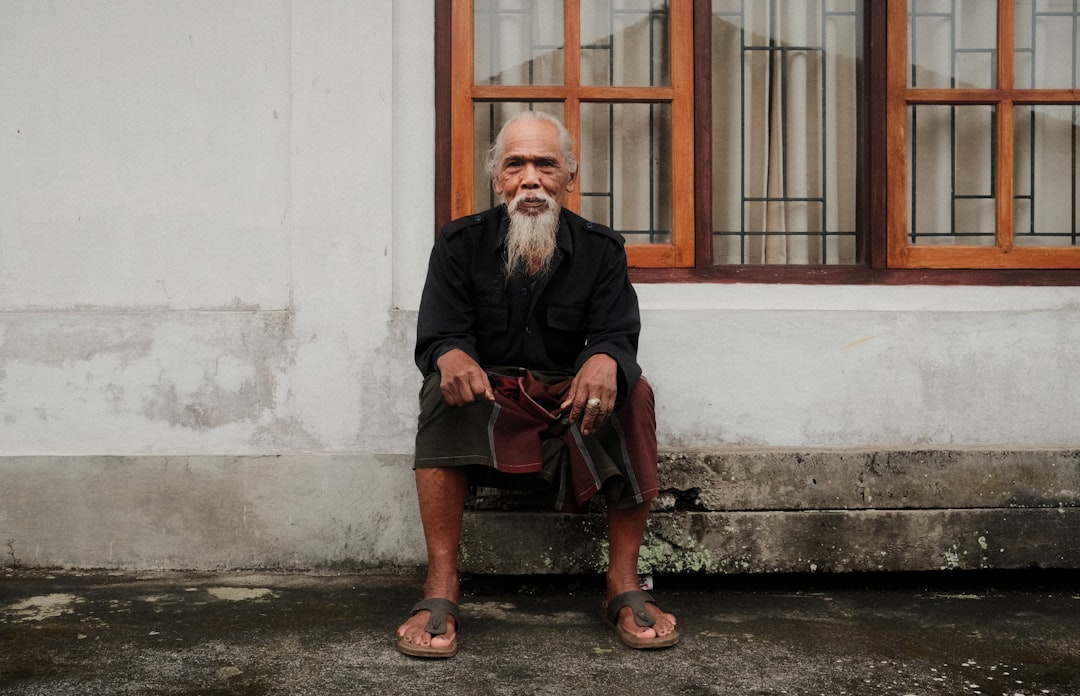 An elderly man sits in front of a window.,