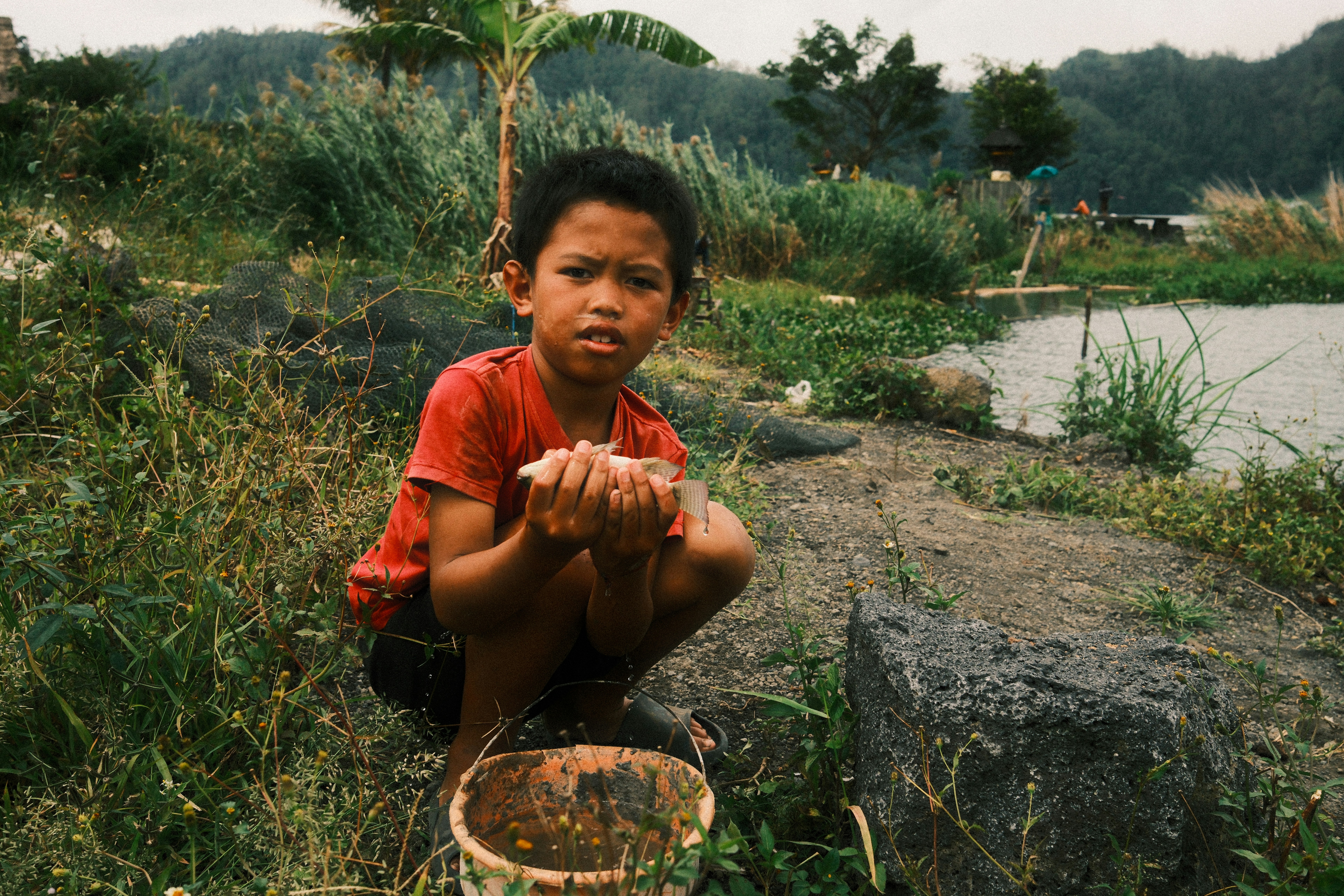 Boy crouches by water, holding a small fish.