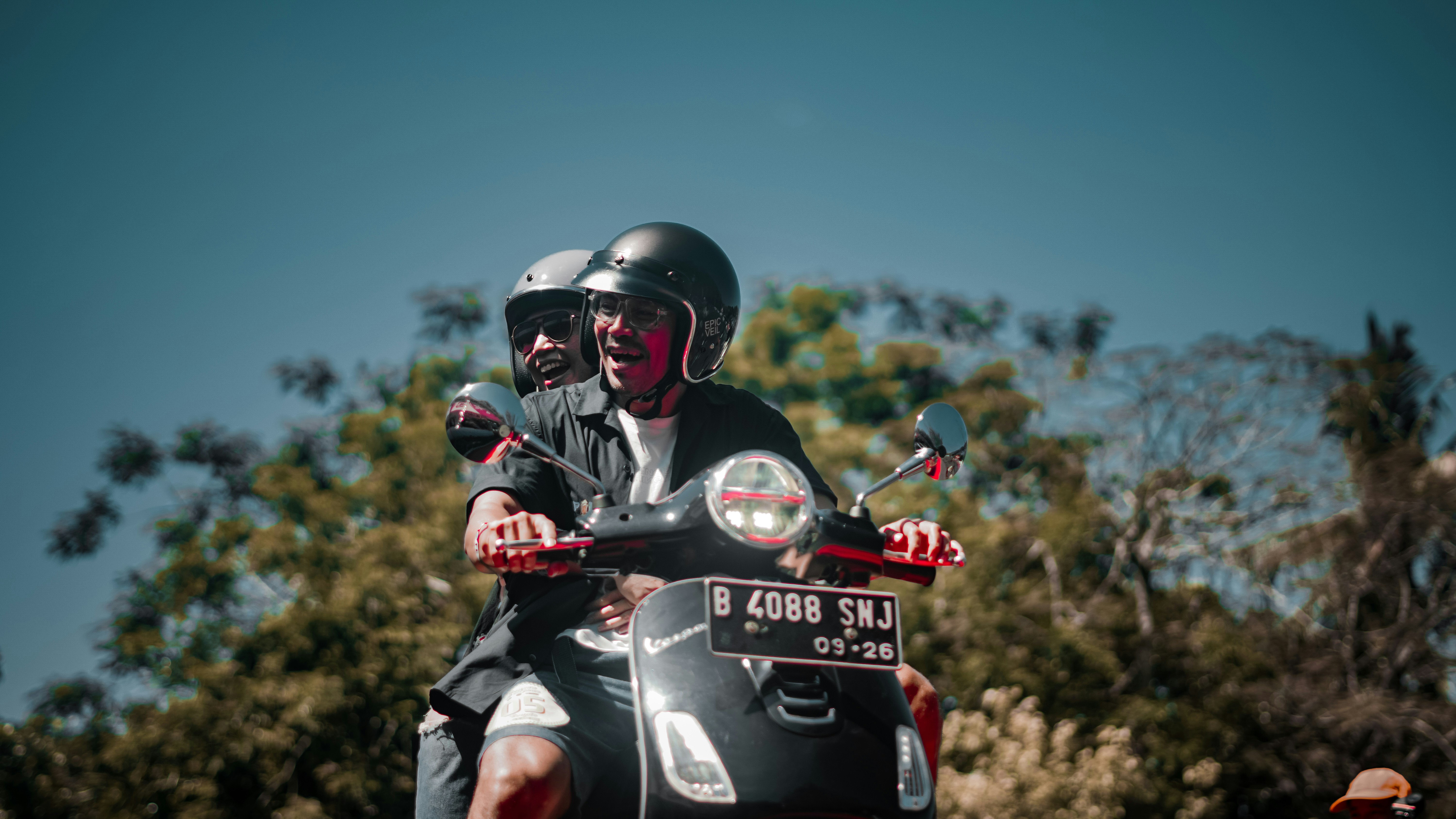 Two people ride a scooter outdoors, smiling.