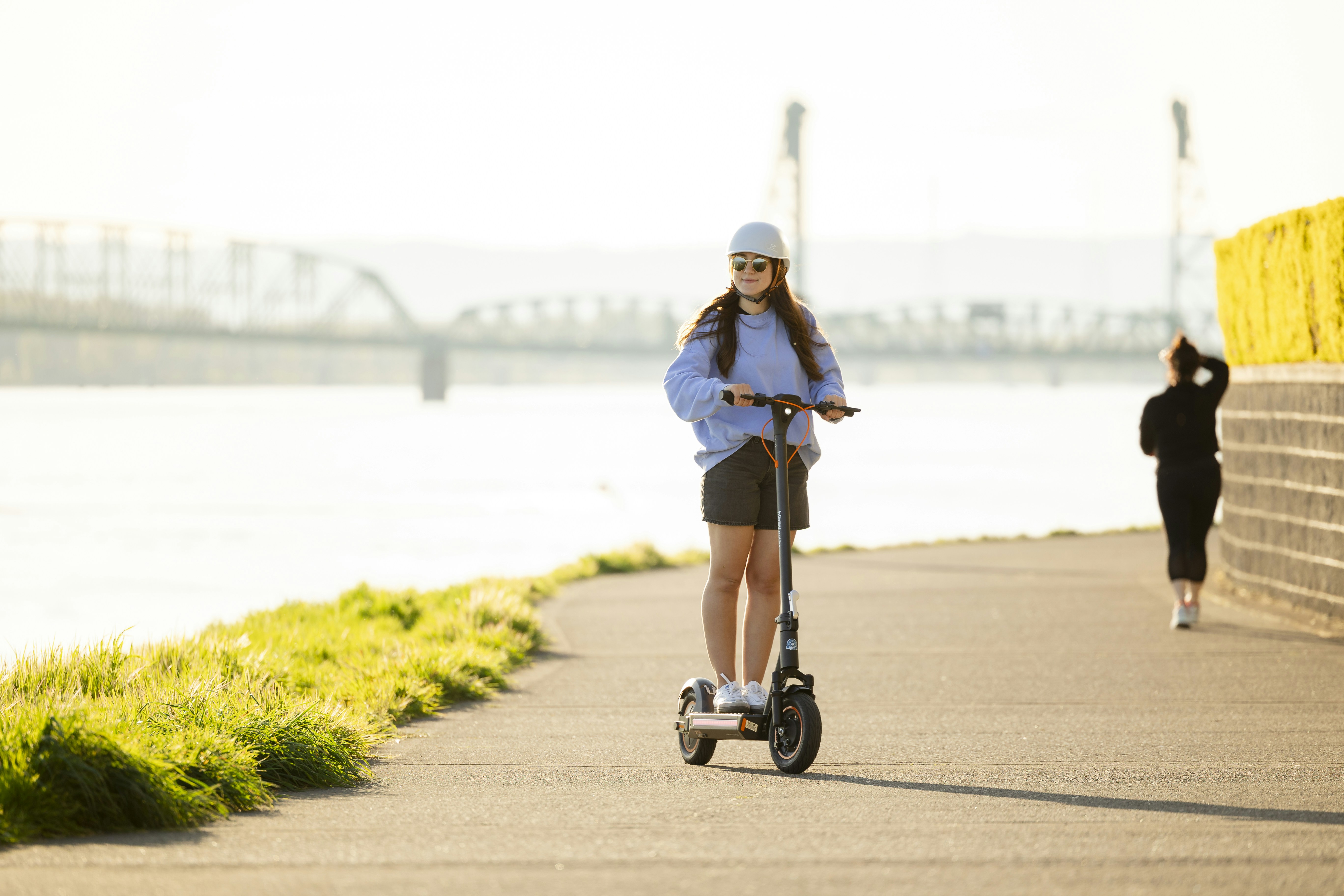 Woman rides a scooter along a waterfront path.