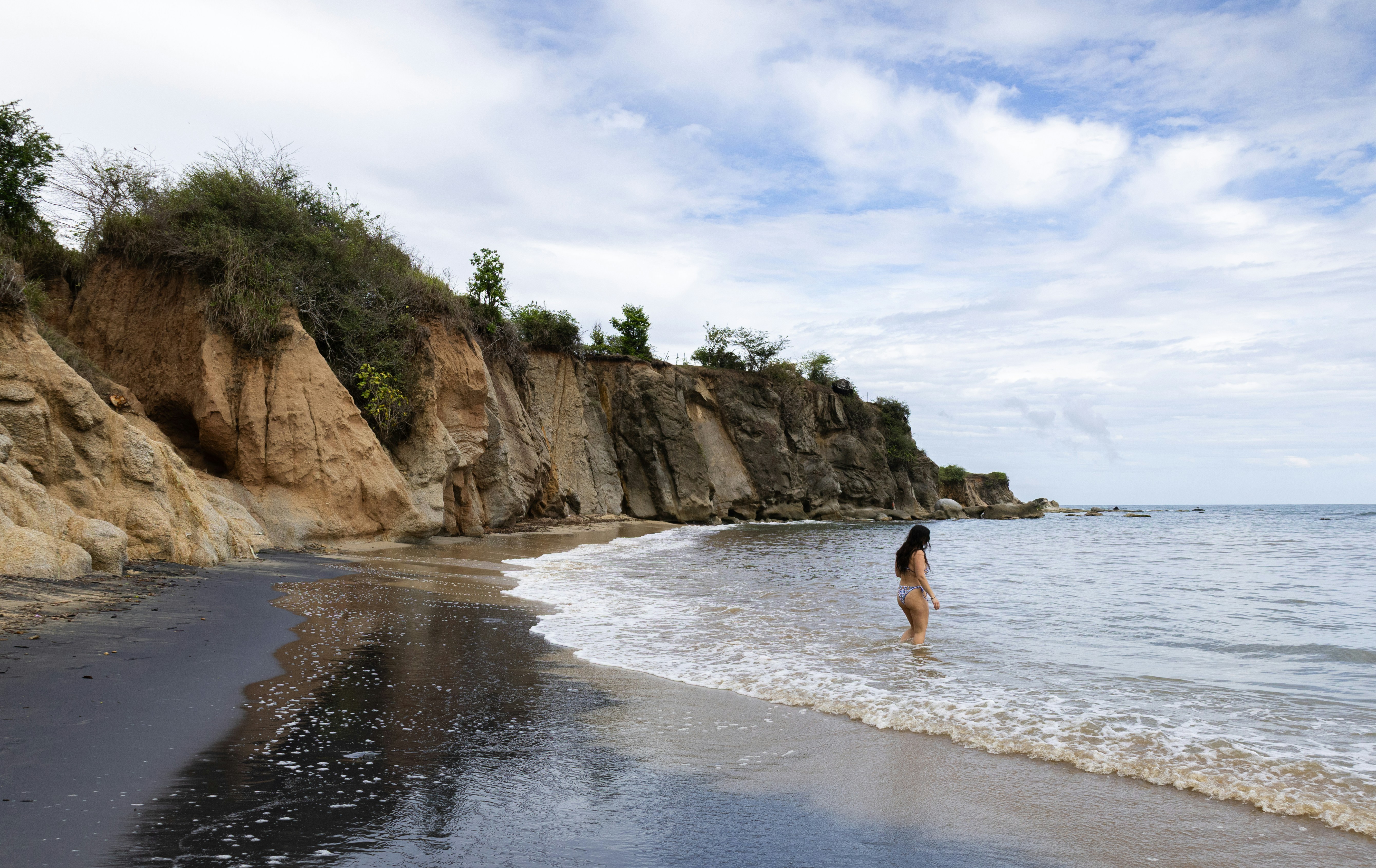 A person stands in the ocean by a cliff.