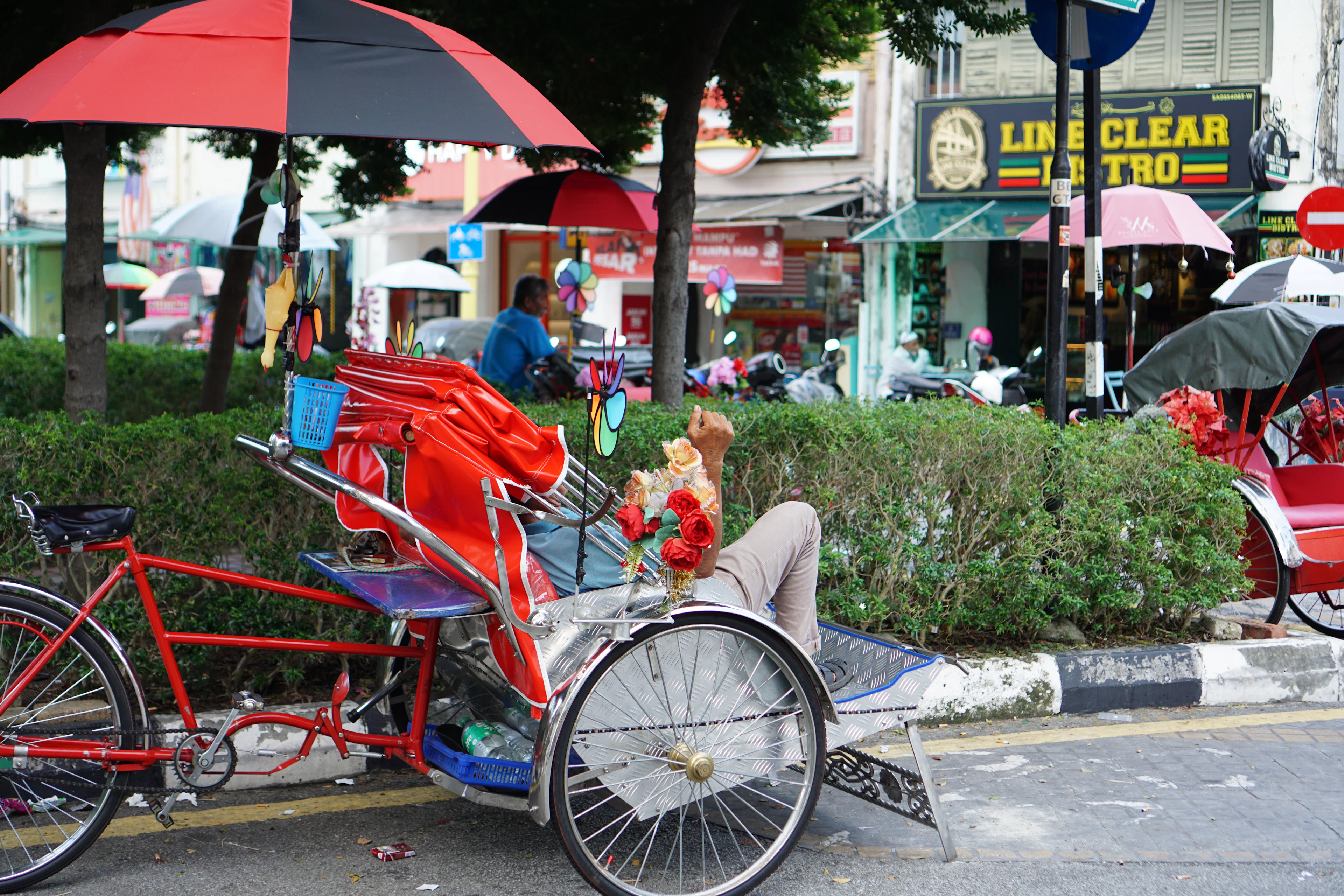 A trishaw rider resting beneath bright umbrellas on a Penang street
