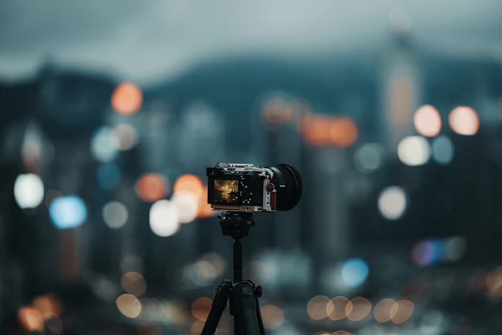 Tripoda kurulu aynasız bir fotoğraf makinesi, mavi saatte ışıkları yanmaya başlamış bir şehir silüetine dönük duruyor