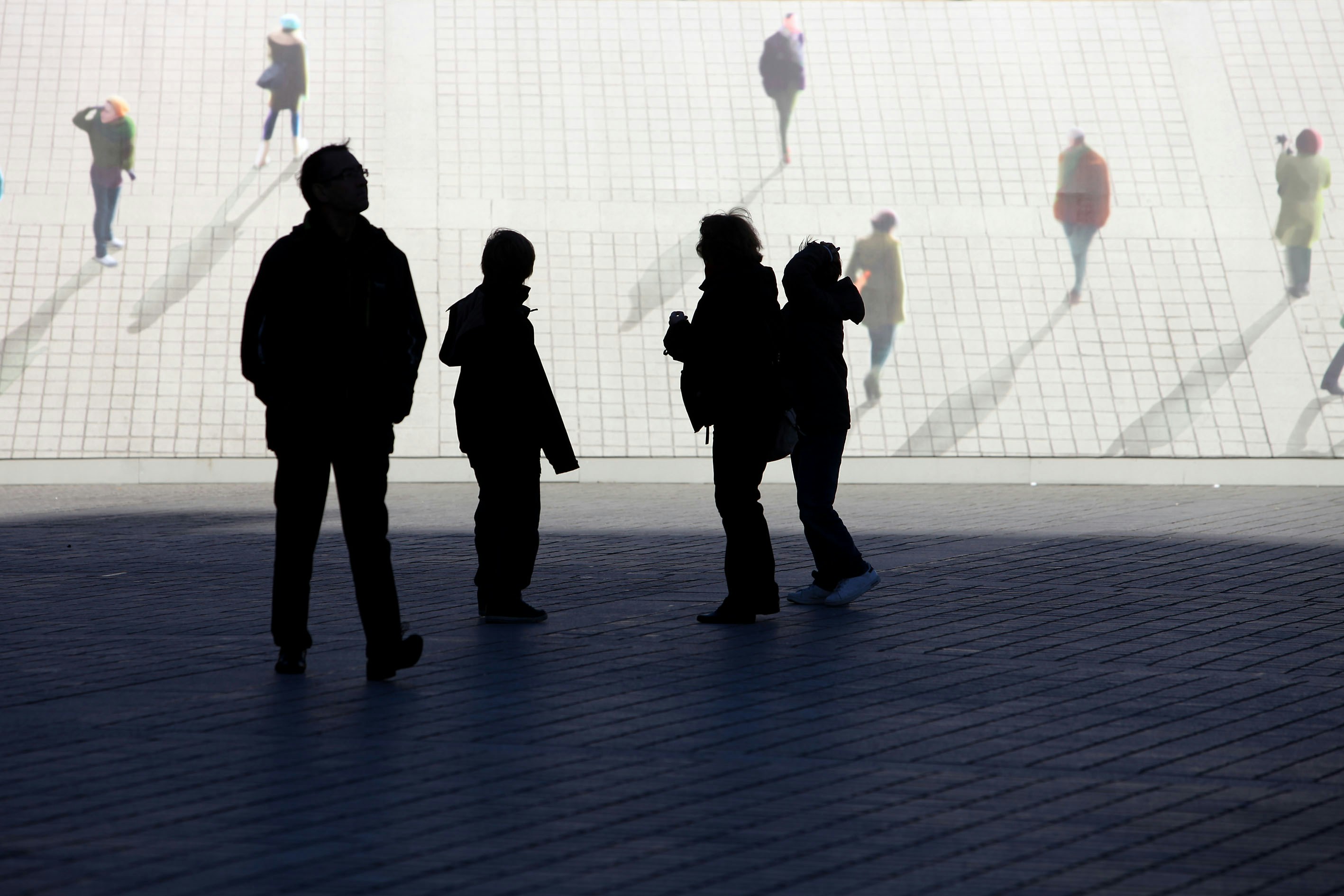 Las siluetas de las personas caminan sobre una zona soleada y pavimentada.