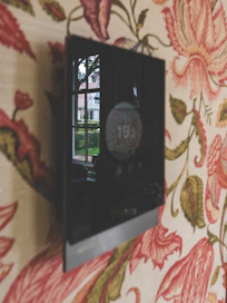 A black, digital thermostat is on a floral wallpapered wall.