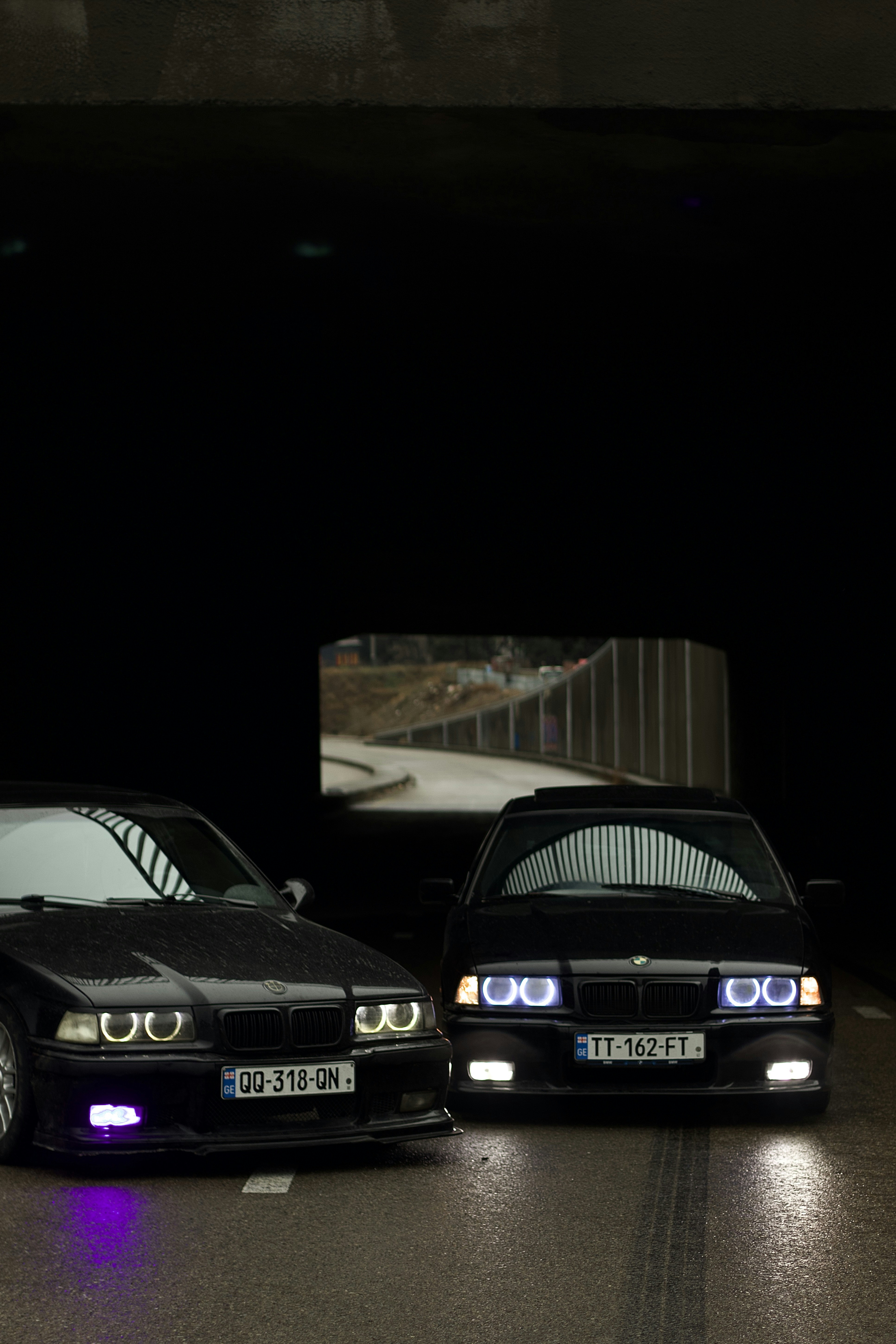 Two black bmws illuminated in a dark tunnel.