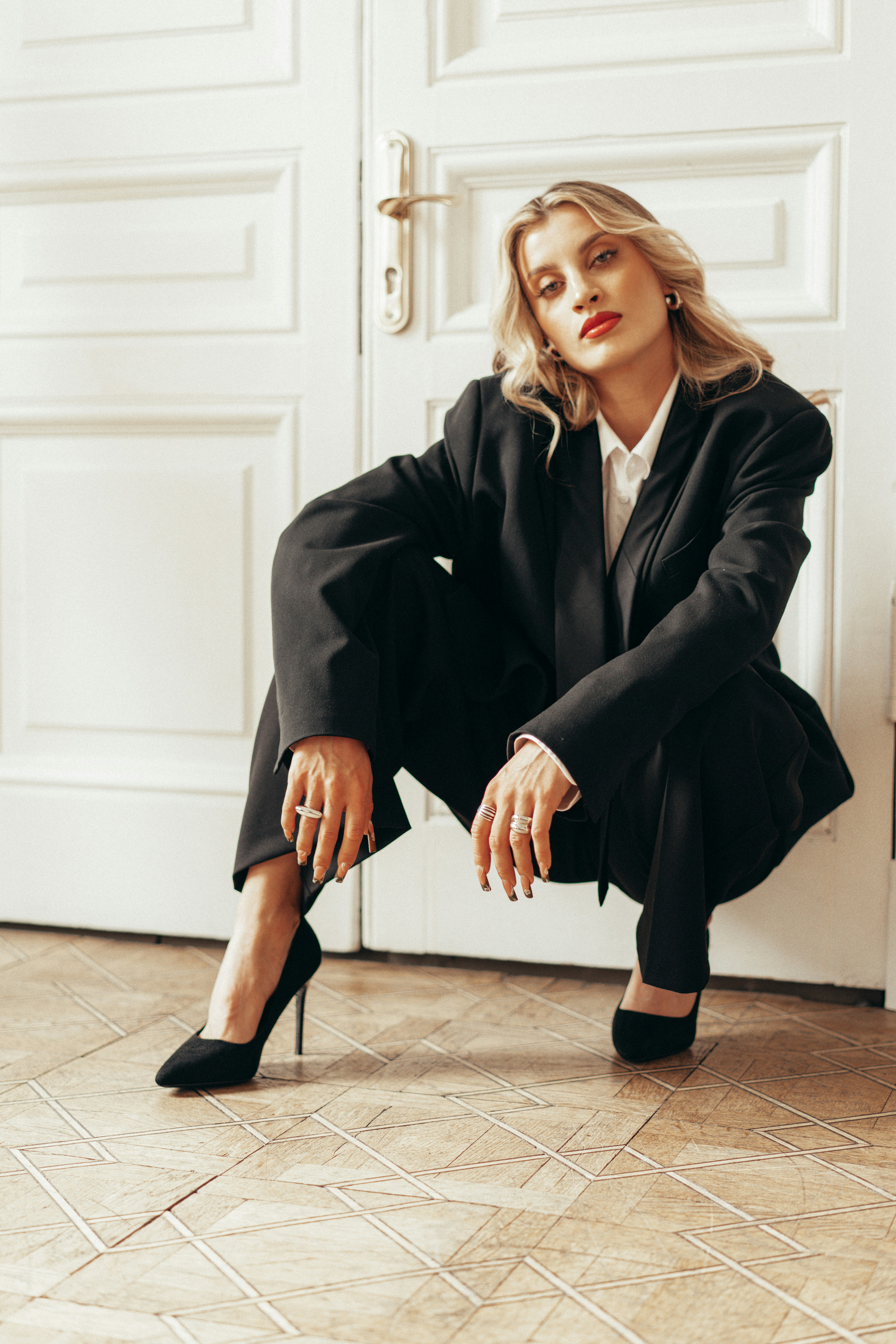Woman in an oversized black suit posing by a white door.