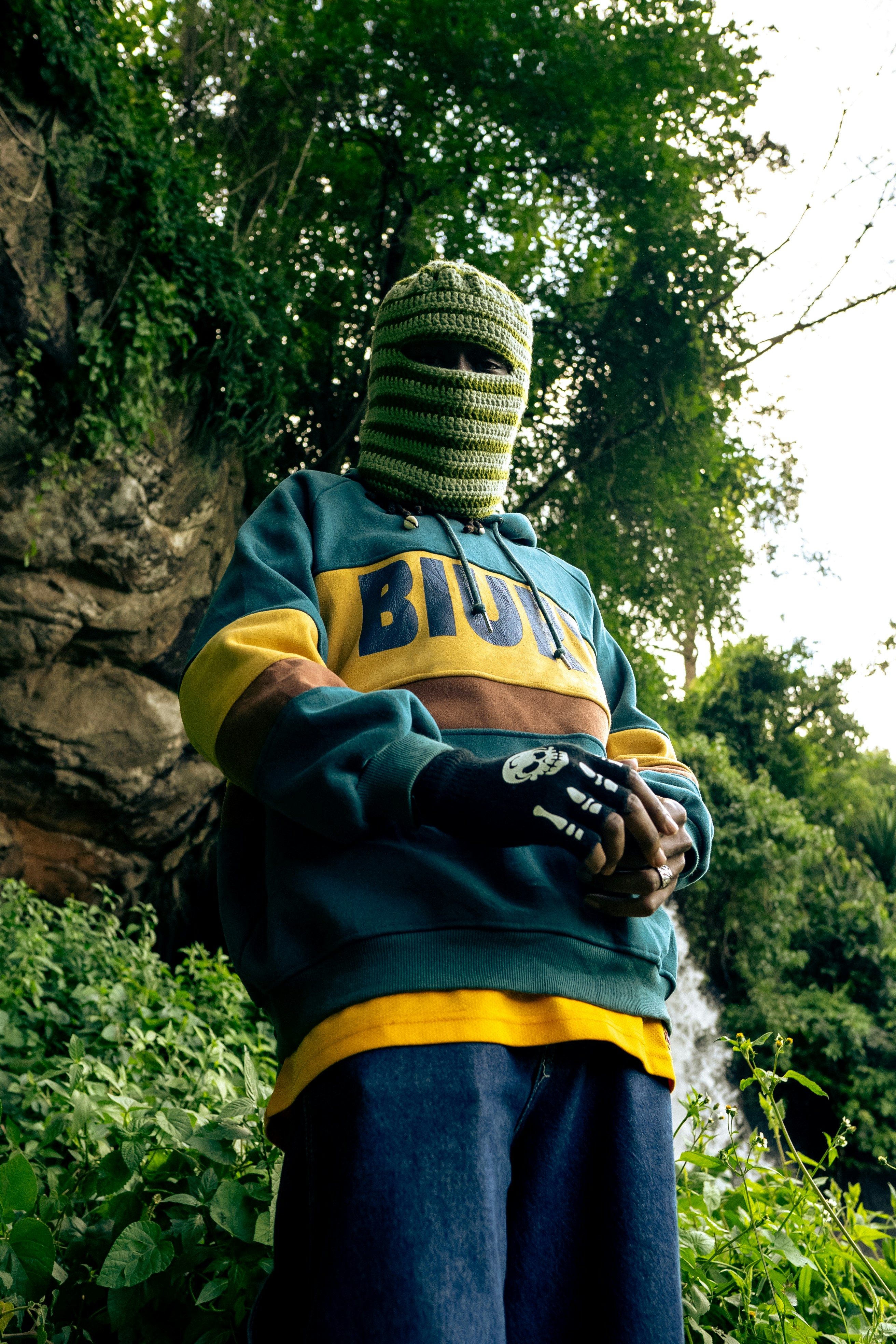 Person in a striped balaclava and hoodie standing in a forest.