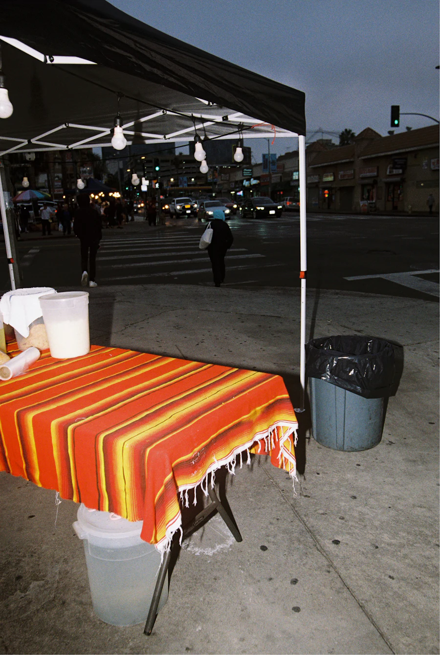 A sidewalk food table under a red and orange striped serape cloth with a jug of horchata, a city intersection glowing behind it at dusk