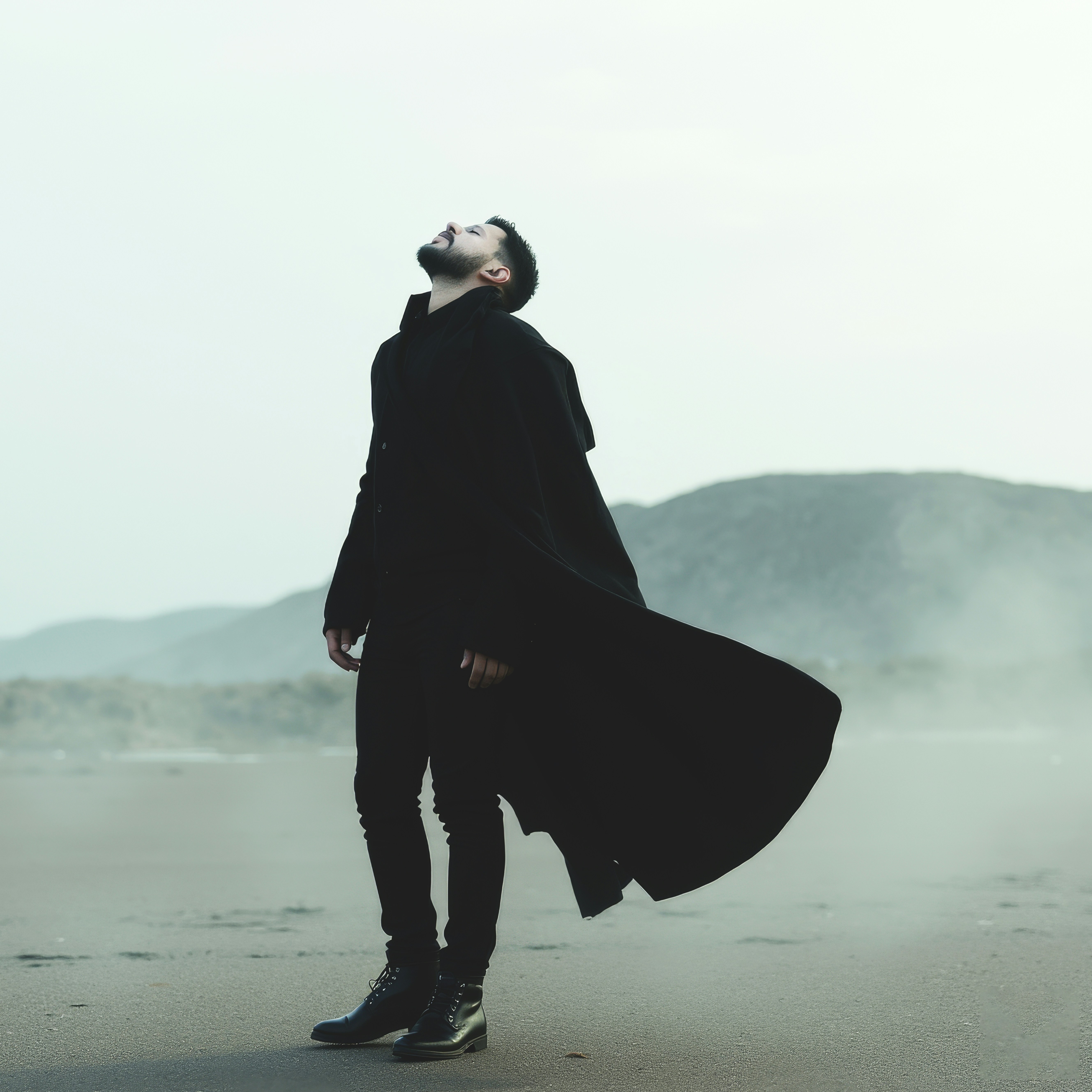 Man looks upwards, wearing a black coat on a beach.