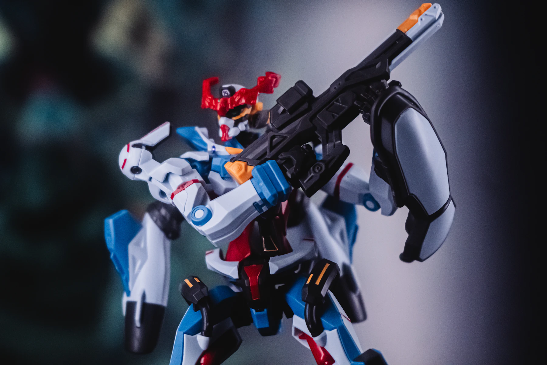 A blue, white and red robot model posed with a black rifle against a dark blurred backdrop