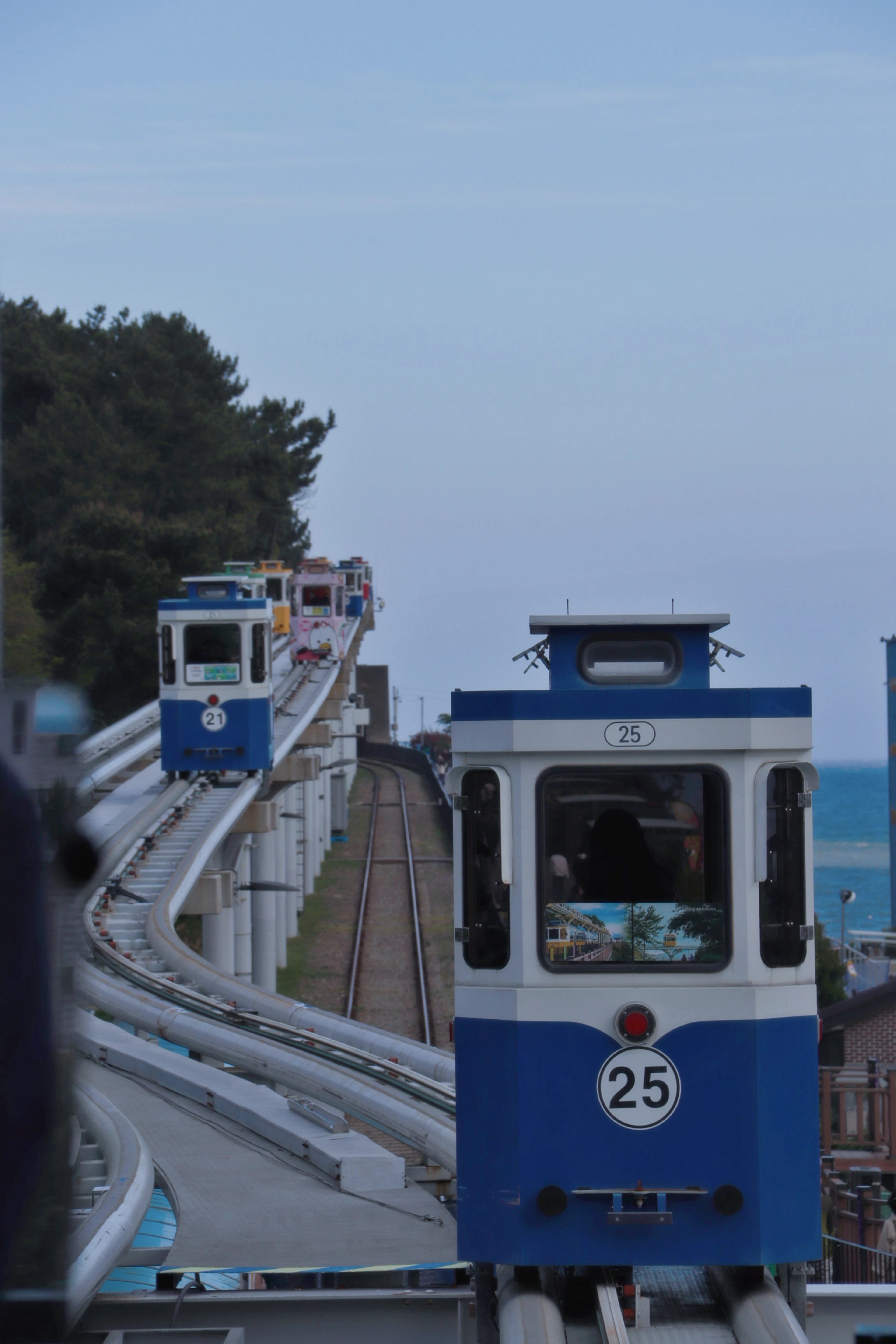 Blue-and-white coastal tram cars ride elevated tracks above the sea near Busan, pine trees along the hillside.
