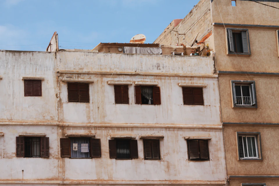 Two-story building with white walls and brown shutters on a street corner with trees