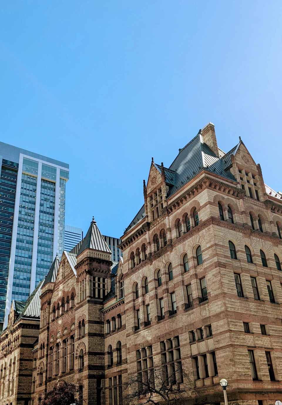 The image depicts a cityscape with two distinct buildings, one large and ornate brick building on the left side and a smaller, modern glass skyscraper on the right side. The brick building features multiple levels and a pointed roof, while the glass skyscraper has a flat top and numerous windows that reflect the surrounding environment.

The sky above is clear blue with no clouds visible, suggesting a bright and sunny day.