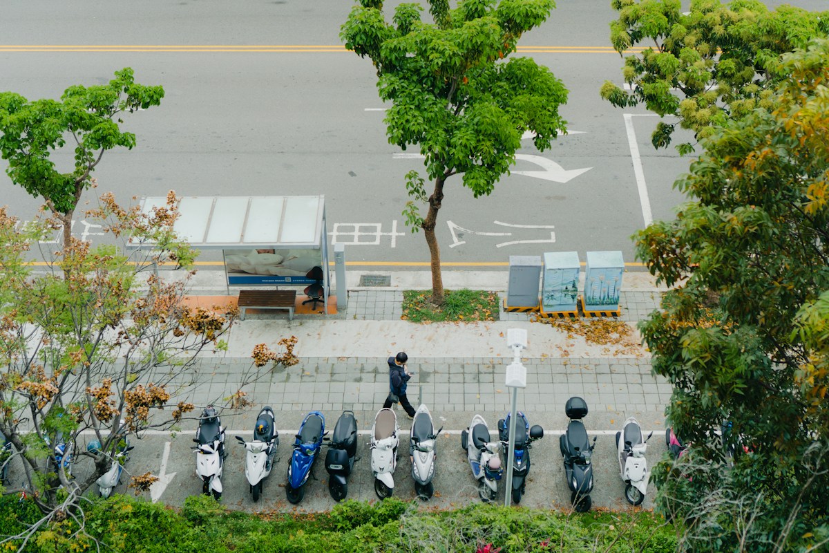 A row of scooters parked along a tree-lined street next to a bus shelter in Taichung, Taiwan, seen from above