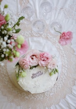 A birthday cake is surrounded by flowers and glassware.