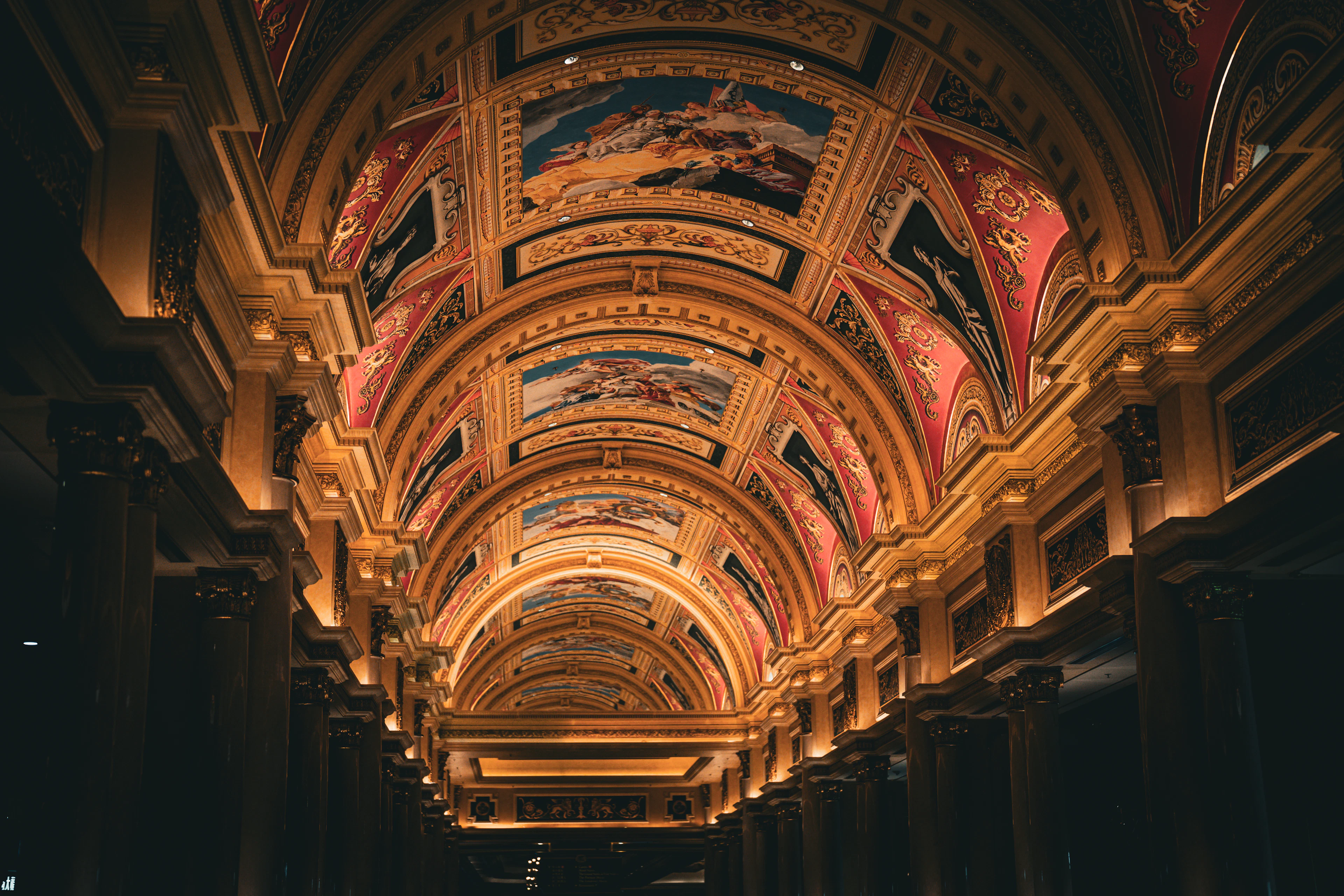 The barrel-vaulted arcade ceiling of the Venetian Macao in Cotai, painted with sky frescoes in gilded frames and lit gold above a receding marble colonnade.