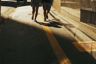 Two people are walking down a sunlit, shaded street.