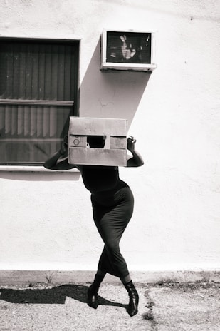 Woman with a box head stands before an outdoor tv.