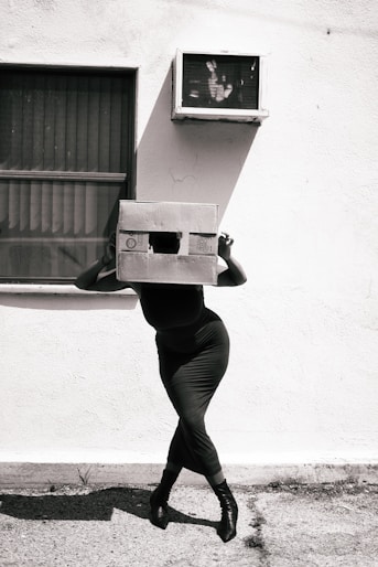 Woman with a box head stands before an outdoor tv.