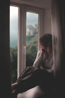 A woman sits by a window, gazing out.